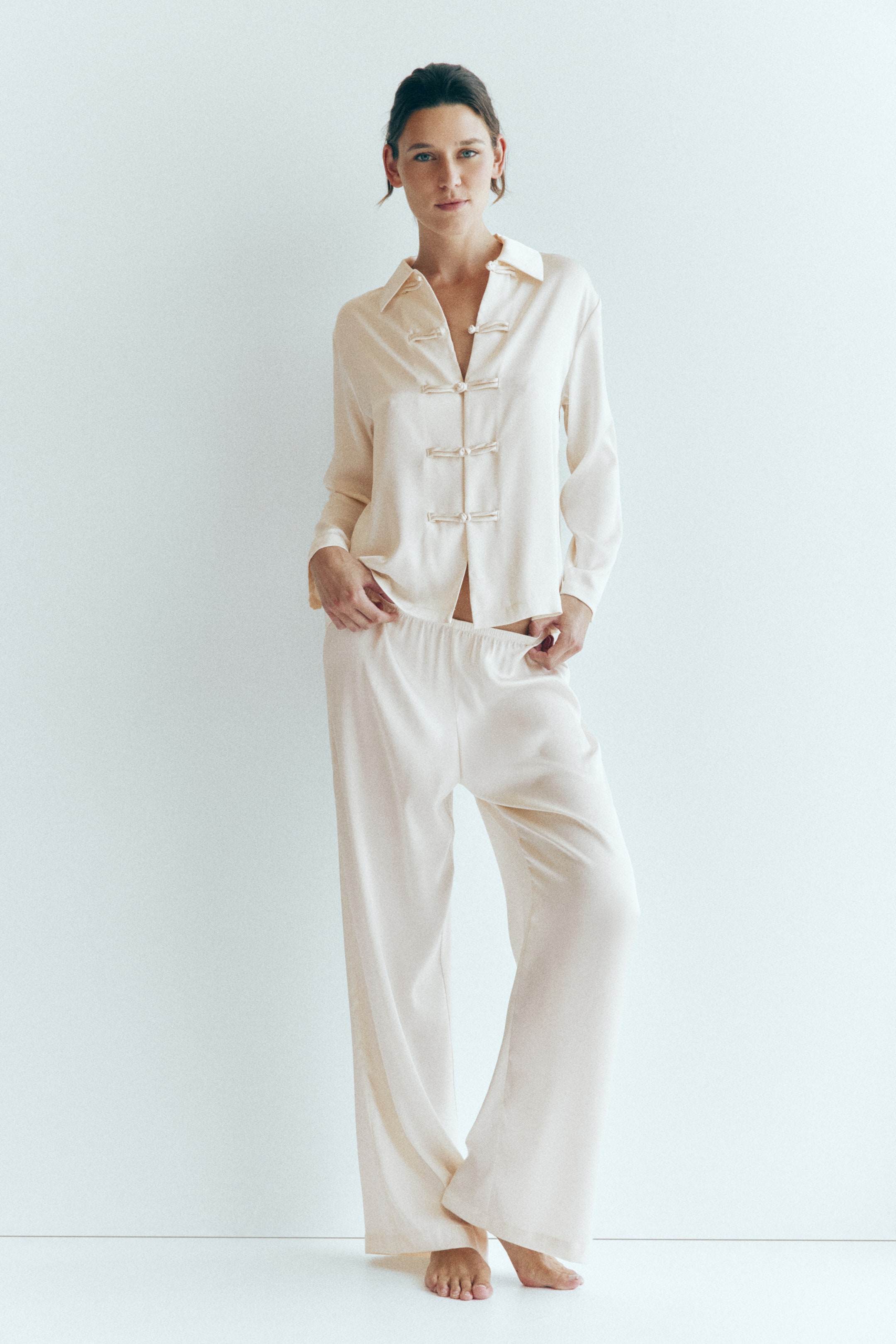 Vezi imaginea mai mare: Un set de pijama din satin, de culoare crem, este purtat de model, alcătuit dintr-o cămașă cu mânecă lungă, cu guler, umeri lăsați și nasturi cu nod în față. Modelul stă în picioare și ține de betelia elastică a pantalonilor asortați, cu croială largă.