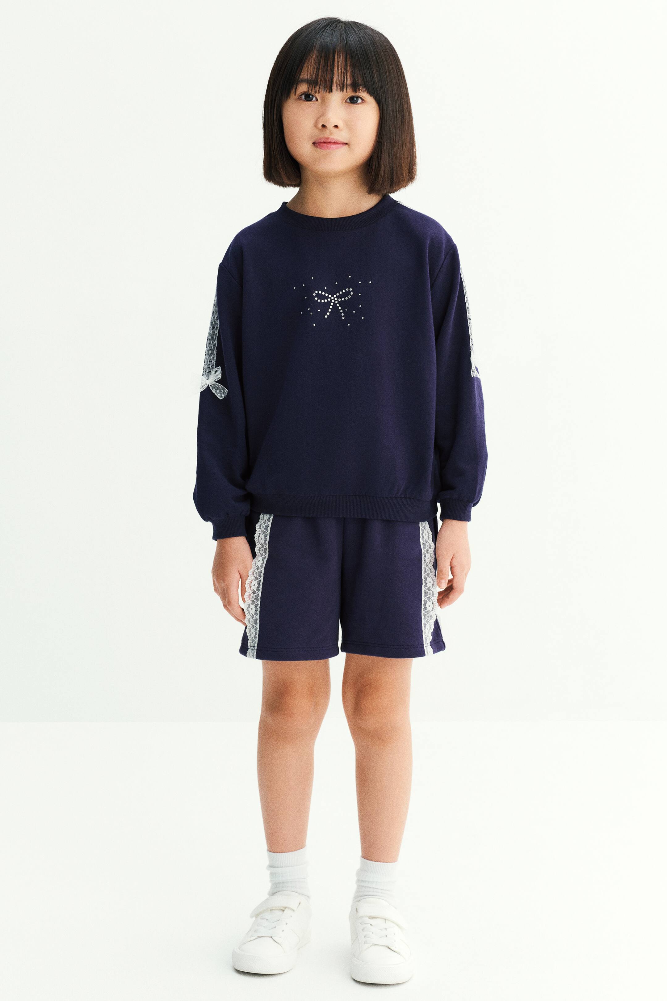 Xem ảnh lớn hơn: Người mẫu đang mặc một bộ đồ gồm áo sweatshirt màu xanh navy và quần short đồng bộ. Áo sweatshirt tay dài nổi bật với họa tiết nơ đính rhinestone ở ngực và các mảng ren trắng có nơ nhỏ dọc theo tay áo, trong khi quần short được nhấn bằng đường viền ren trắng chạy dọc hai bên.