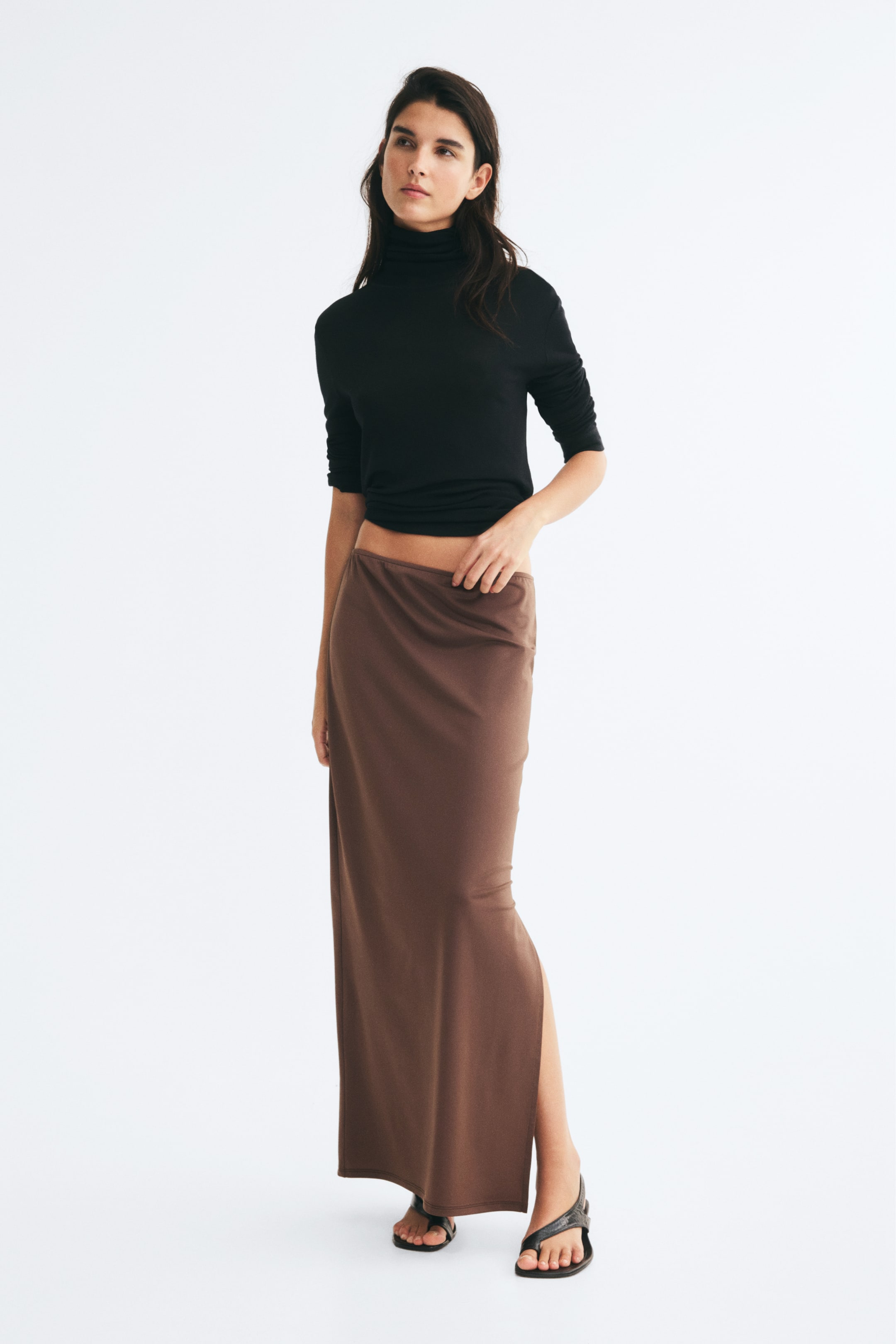 Visa större bild: En lång, figursydd brun kjol i stretchjersey bärs av modellen och har en elastisk midja samt en rak fåll med en slits i sidan. Modellen rör lätt vid kjolens linning, som matchas med en långärmad svart polotröja och svarta sandaler.