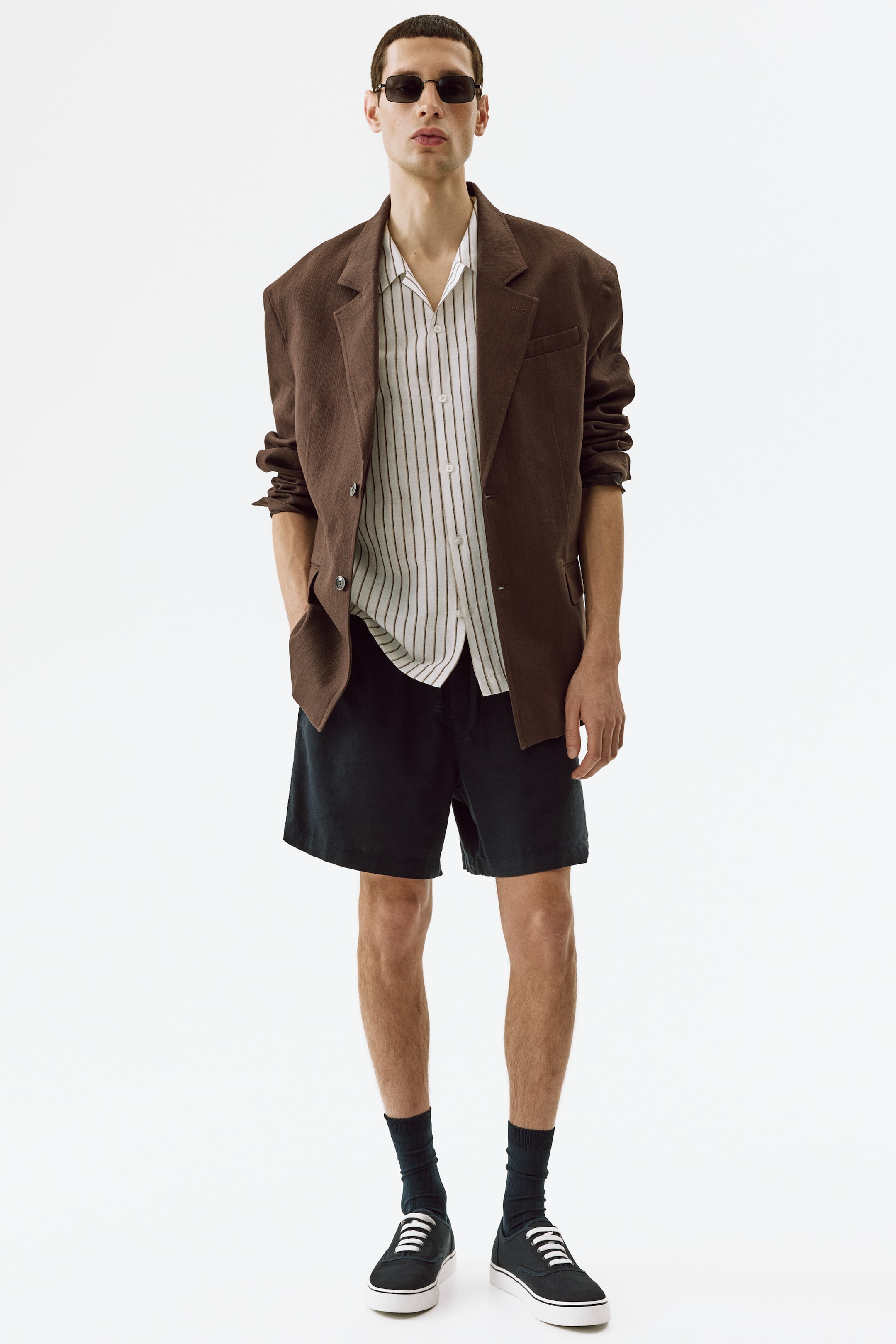 Ingrandisci l'immagine: Un blazer marrone oversize con revers a lancia è indossato sopra una camicia a righe bianche a maniche corte con colletto aperto. Pantaloncini scuri a vestibilità rilassata sono abbinati a calzini scuri al polpaccio, sneakers in tela nera con lacci bianchi e occhiali da sole rettangolari per completare l'ensemble.
