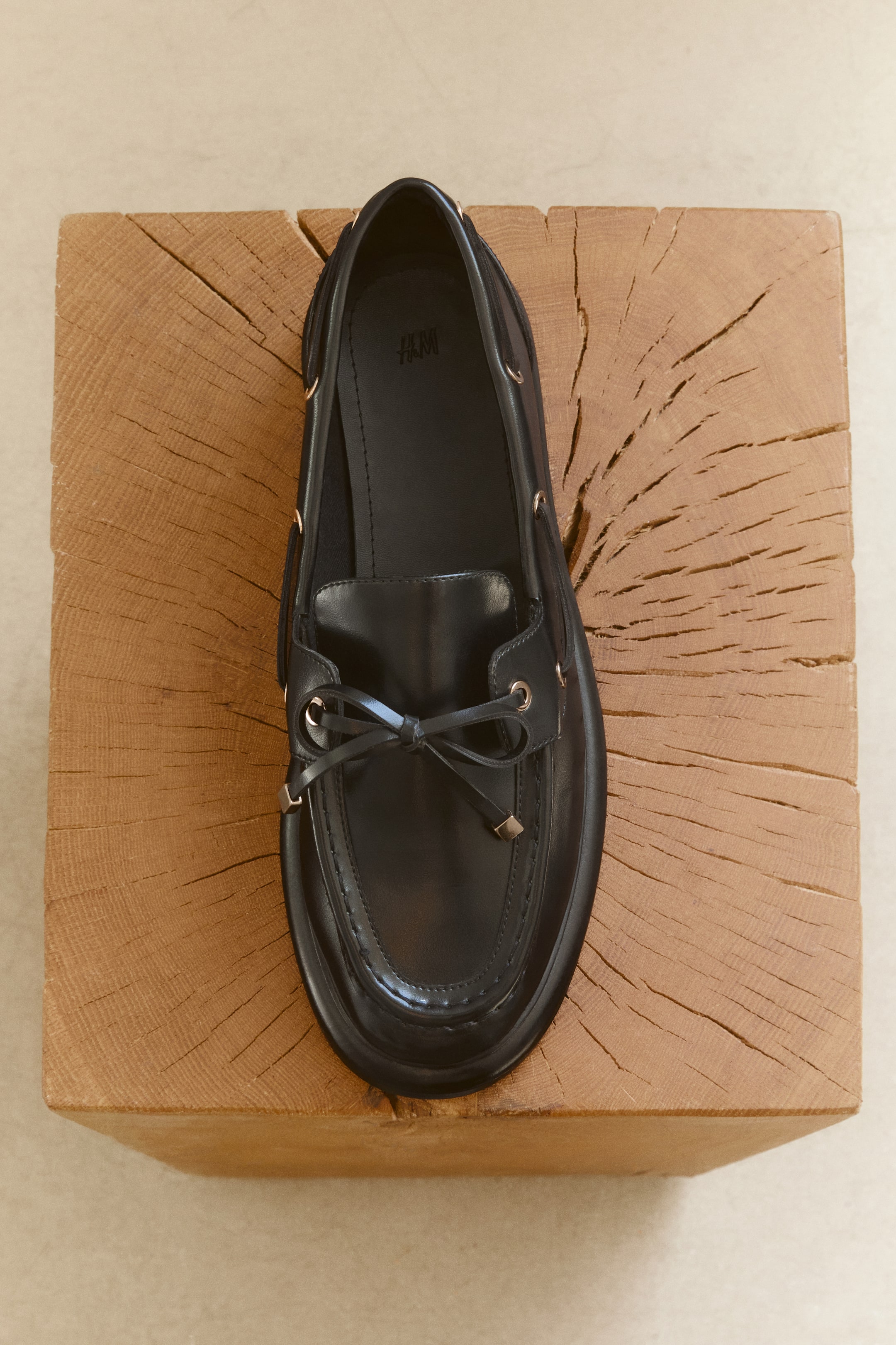 Ver imagen más grande: Una vista cenital muestra un mocasín negro inspirado en los zapatos náuticos, con puntera redondeada y una costura tipo mocasín en relieve. Un cordón decorativo atraviesa ojales metálicos a lo largo de los laterales y se anuda en forma de lazo sobre el empeine delantero.