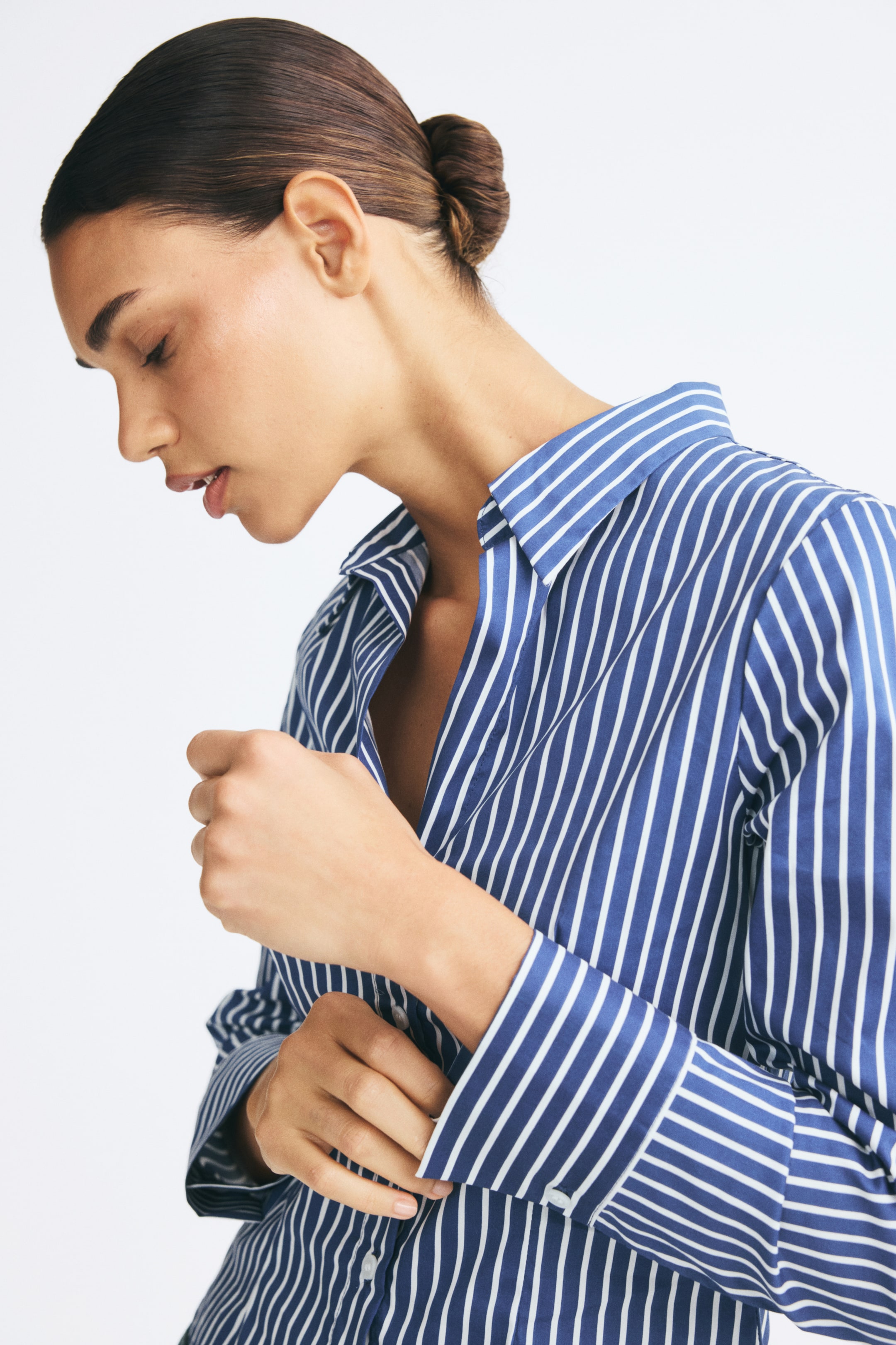 View larger image: A blue and white vertically striped fitted long-sleeve shirt with an open collar is being worn by the model. The model is looking down while adjusting the buttoned cuff of the sleeve.
