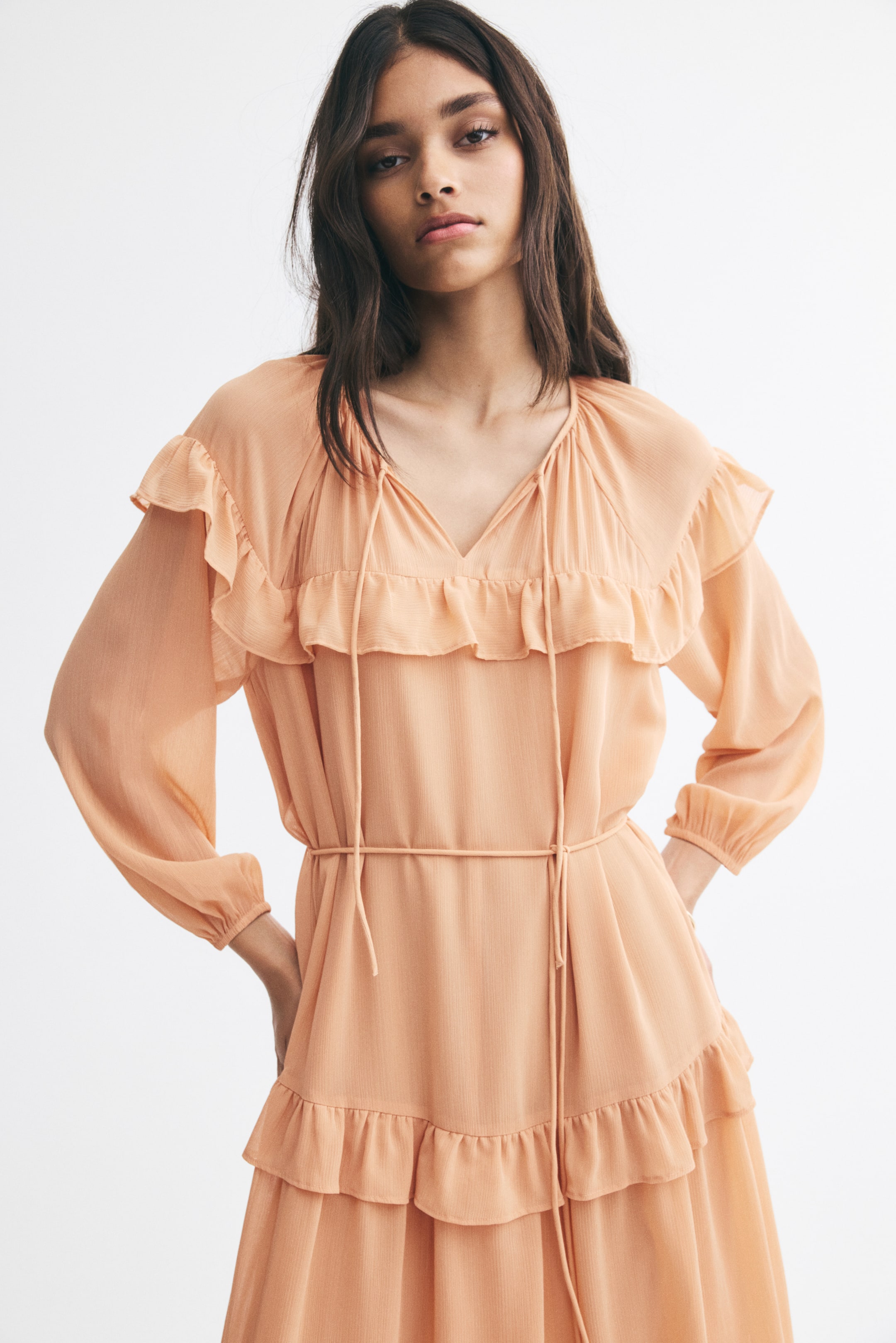 View larger image: The model is wearing a peach-coloured dress in a textured weave, featuring a V-shaped neckline with slender tie details, ruffle trims across the chest and hips, and long balloon sleeves. The dress is cinched at the waist with a thin tie belt as the model poses with her hands on her hips.