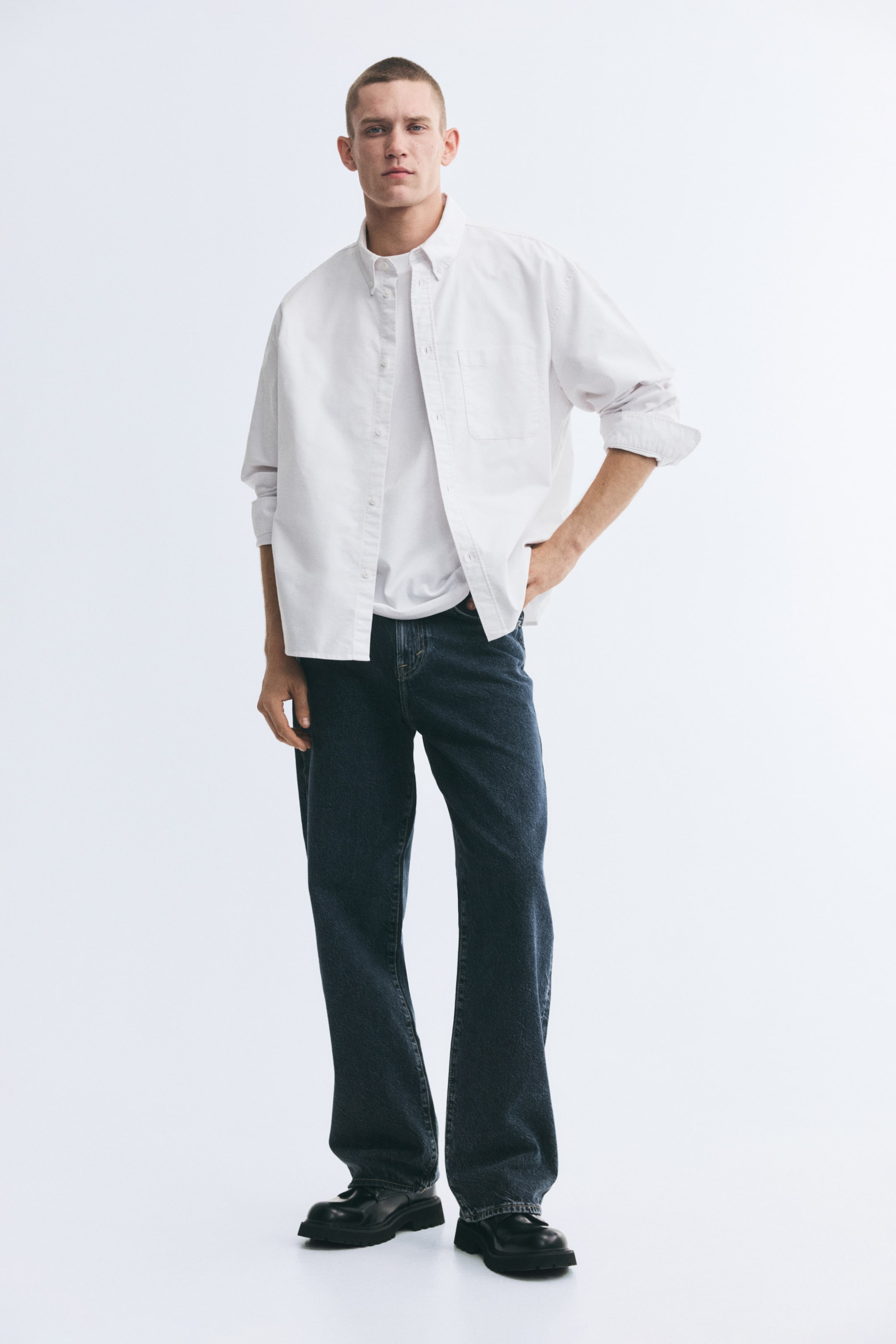 Vis større bilde: Mørkeblå denimjeans med en avslappet, rett passform er båret av modellen, som står med den ene hånden hvilende på hoften. Jeansene er stylet med en hvit T-skjorte under en åpen hvit skjorte med knapper og kombinert med svarte sko.