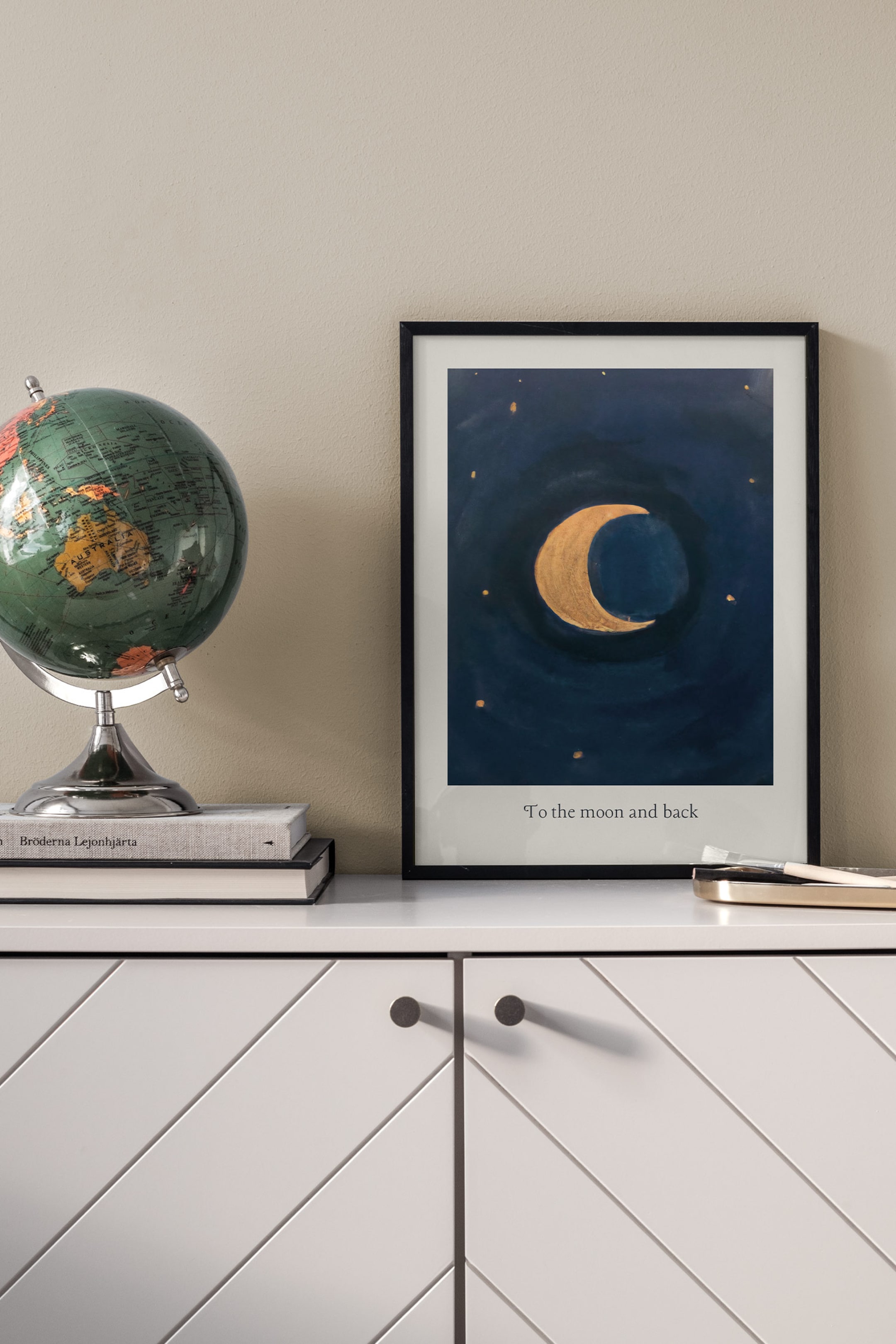 Grotere afbeelding bekijken: Een rechthoekige art print, omlijst door een dun zwart frame, is recht van voren gefotografeerd en toont een getextureerde gouden halve maan omringd door kleine gouden sterren op een donker marineblauwe achtergrond. De zin "To the moon and back" staat in een zwart serif-lettertype binnen de witte rand aan de onderkant van de print.