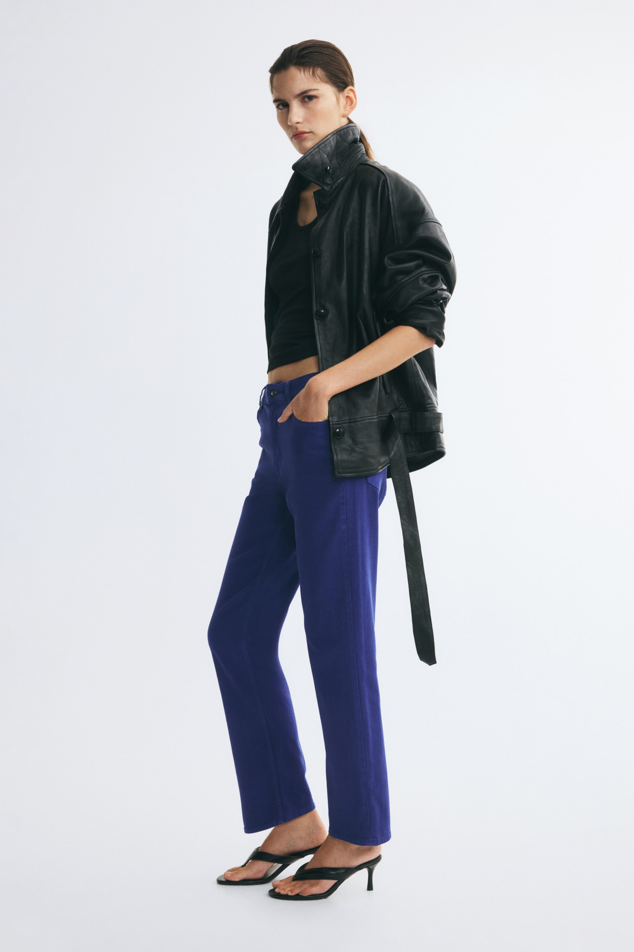 Visualizza immagine più grande: Pantaloni in twill viola a vita alta con gamba slim, indossati dalla modella in posa con una mano appoggiata nella tasca anteriore. I pantaloni sono abbinati a un crop top nero, una giacca di pelle nera e sandali neri con tacco.