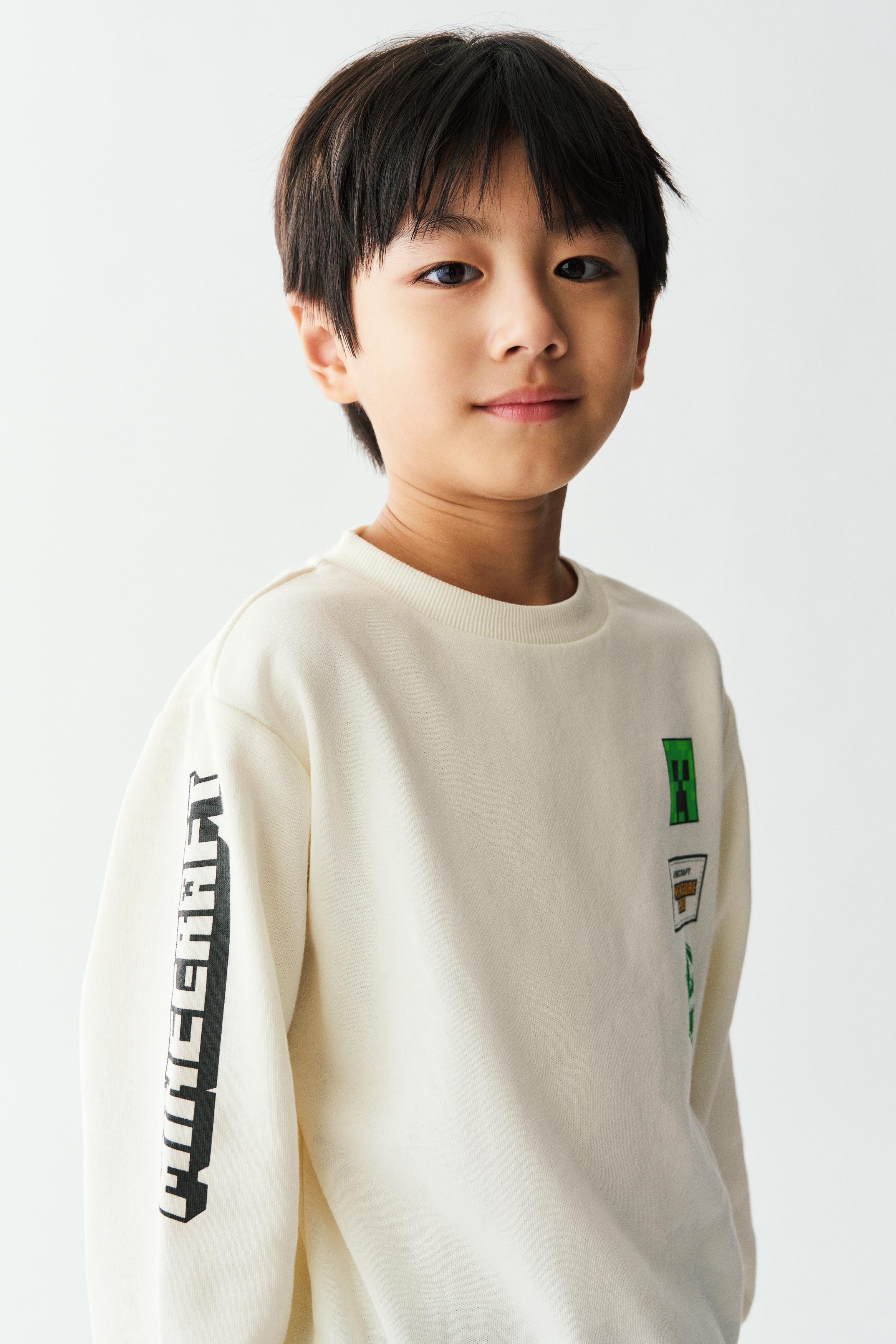 Má»t chiáº¿c Ã¡o sweatshirt mÃ u tráº¯ng ngÃ  ÄÆ°á»£c chá»¥p tá»« phÃ­a trÆ°á»c, ná»i báº­t vá»i cá» trÃ²n bo rib, vai xuÃ´i vÃ  bo gáº¥u cÃ¹ng cá» tay rá»ng. Pháº§n ngá»±c cÃ³ ba hÃ¬nh in chá»§ Äá» Minecraft ÄÆ°á»£c xáº¿p chá»ng theo chiá»u dá»c, trong khi tay Ã¡o pháº£i cÃ³ dÃ²ng chá»¯ mÃ u Äen dáº¡ng pixel cháº¡y dá»c theo chiá»u dÃ i tay Ã¡o.