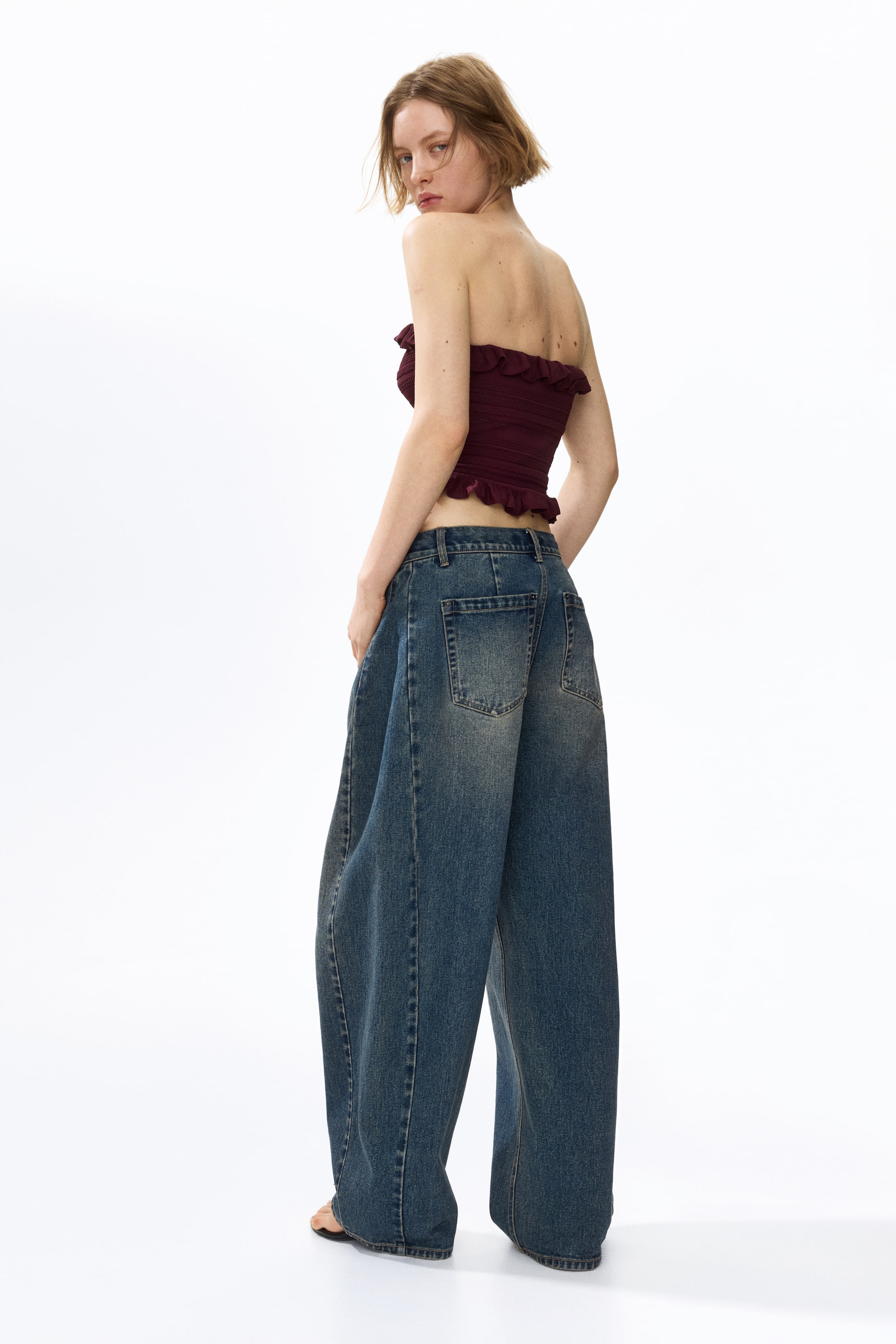 View larger image: The model is wearing a short, fitted bandeau top in a dark red pointelle-knit. It boasts ruffles along its top and bottom edges and is paired with wide-leg jeans.