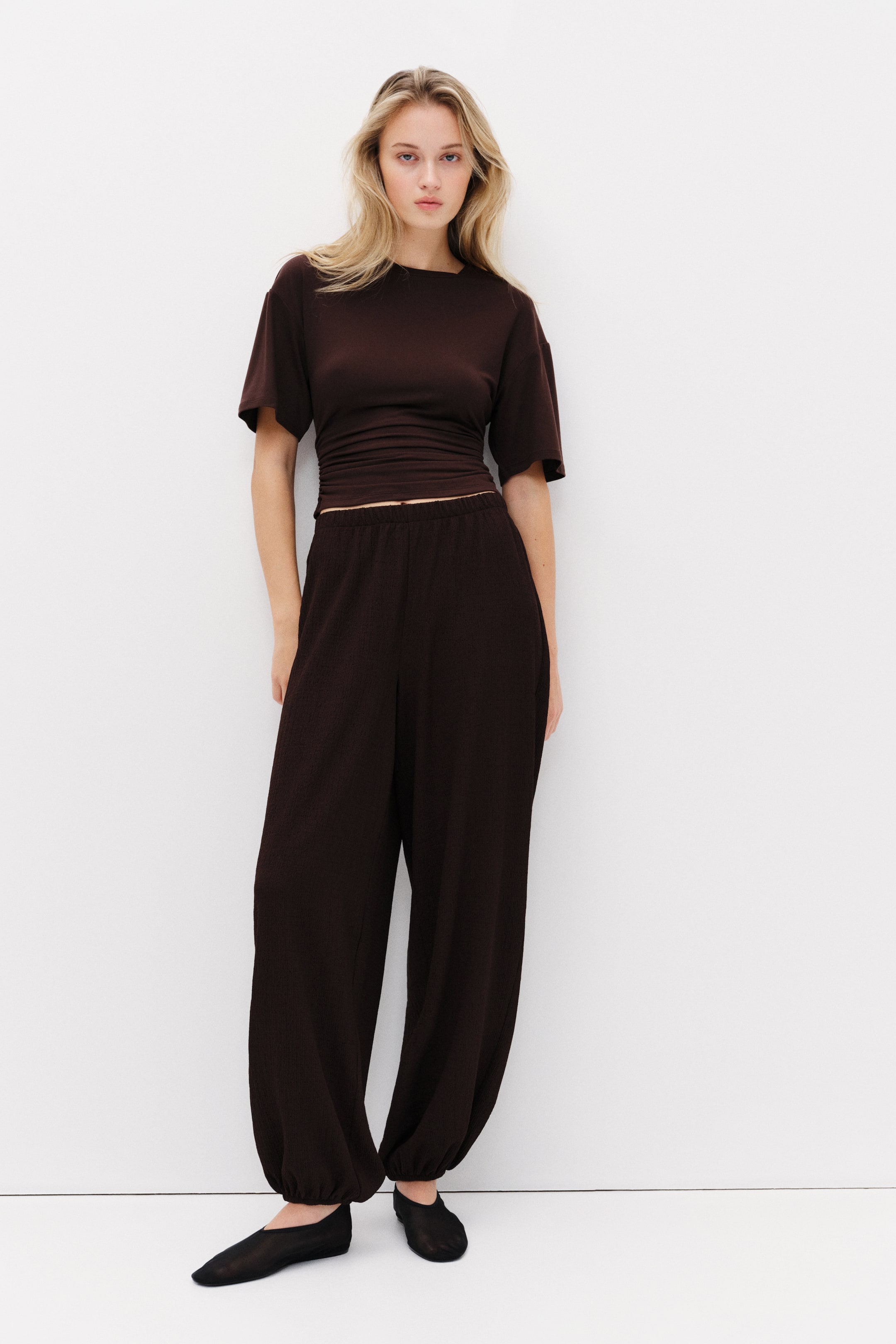 View larger image: "呢件深啡色短袖上衣設有圓領及皺褶腰部，而配搭的闊腳褲則展示彈性腰帶及收腳設計，整體以有質感的布料製成。"