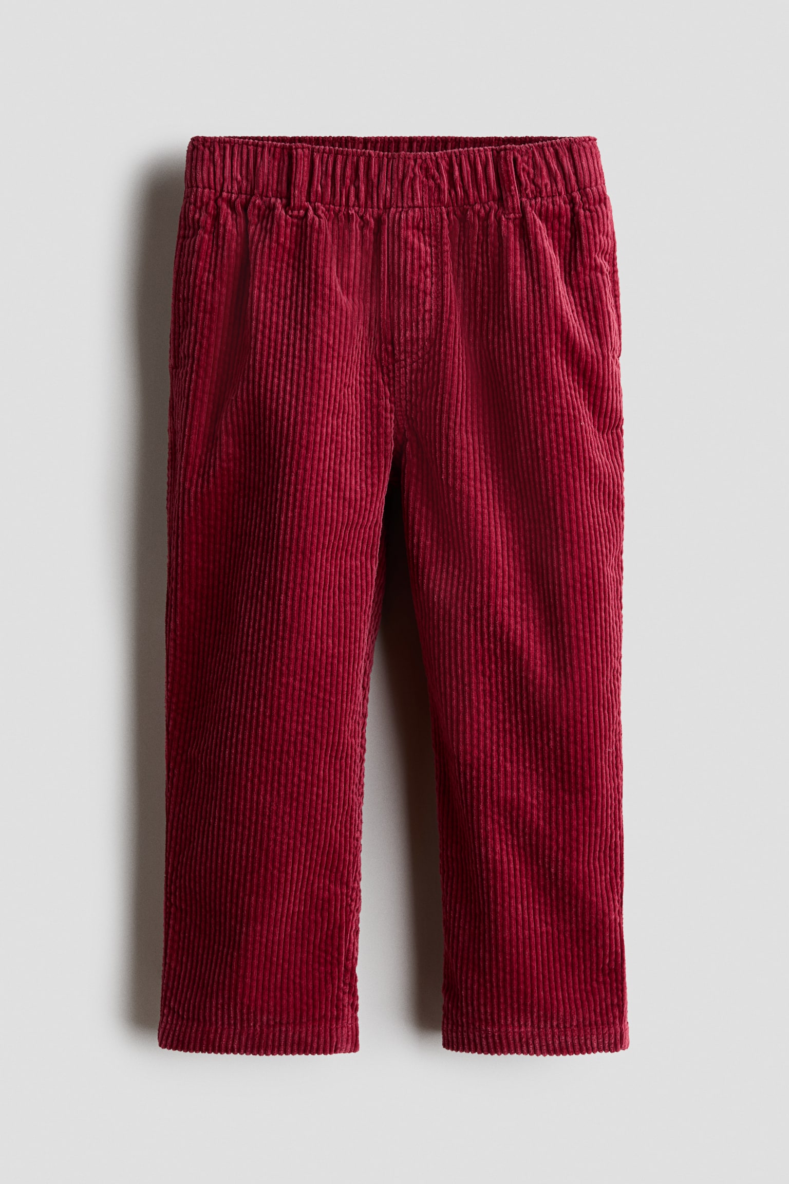 Frontvisning av H&M Adorables-buksen i dyp rÃ¸d kordflÃ¸yel, med samlet elastisk linning og falsk gylf. Buksen med rette ben viser diskrete sidelommer i sÃ¸mmen og en vertikalt ribbet tekstur.