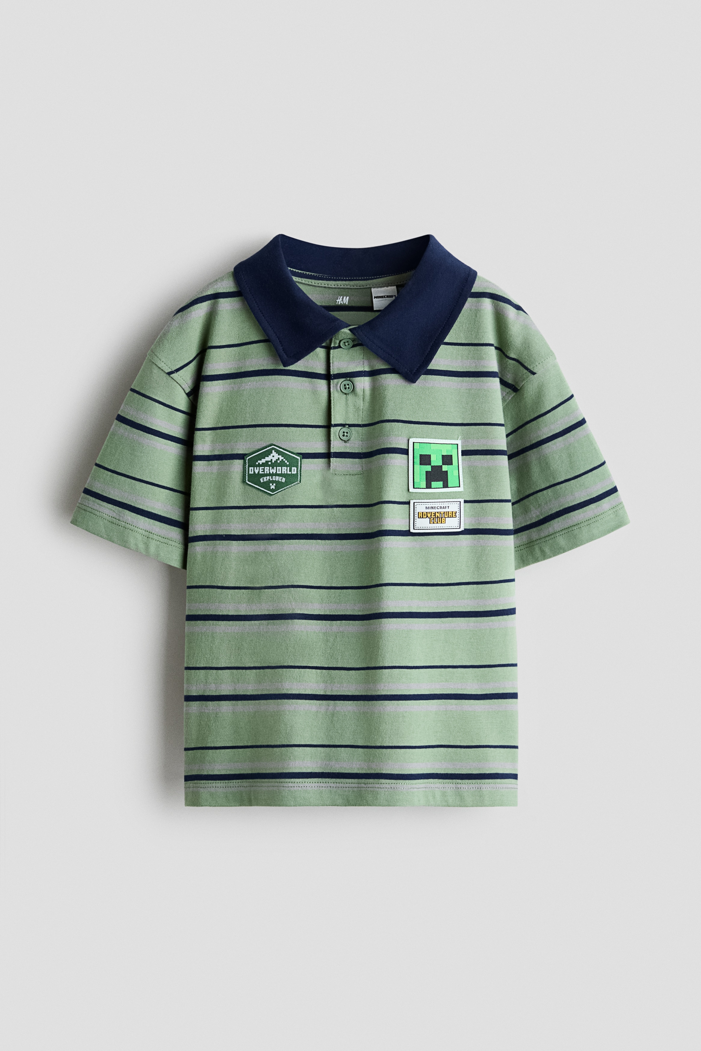 NgÆ°á»i máº«u Äang máº·c má»t chiáº¿c Ã¡o polo dÃ¡ng boxy, káº» sá»c xanh lÃ¡, vá»i cá» báº» mÃ u xanh navy Äáº­m, hÃ ng cÃºc phÃ­a trÆ°á»c vÃ  cÃ¡c miáº¿ng patch trang trÃ­ theo chá»§ Äá» Minecraft á» máº·t trÆ°á»c. NgÆ°á»i máº«u cÅ©ng Äang Äá»i má»t chiáº¿c mÅ© lÆ°á»¡i trai mÃ u xanh lÃ¡ Äá»ng bá» vÃ  Äang cáº§m, chá»nh láº¡i mÅ© trÃªn Äáº§u.