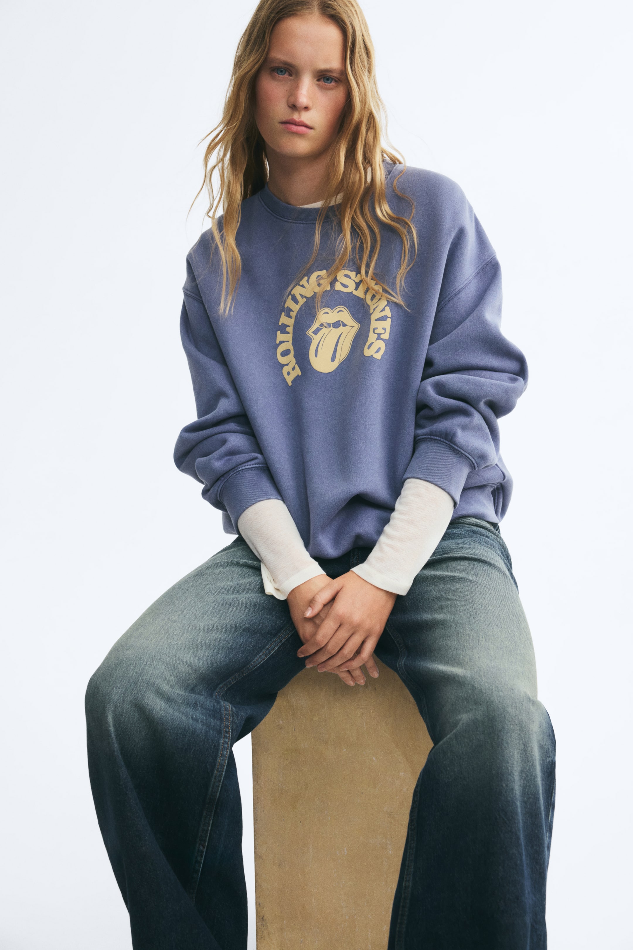 Zobrazit větší obrázek: Modelka má na sobě oversized modrou mikinu s žebrovaným kulatým výstřihem, spuštěnými rameny a žlutým grafickým potiskem loga „Rolling Stones“. Mikina je vrstvena přes krémový top s dlouhým rukávem a doplněna širokými modrými džíny.