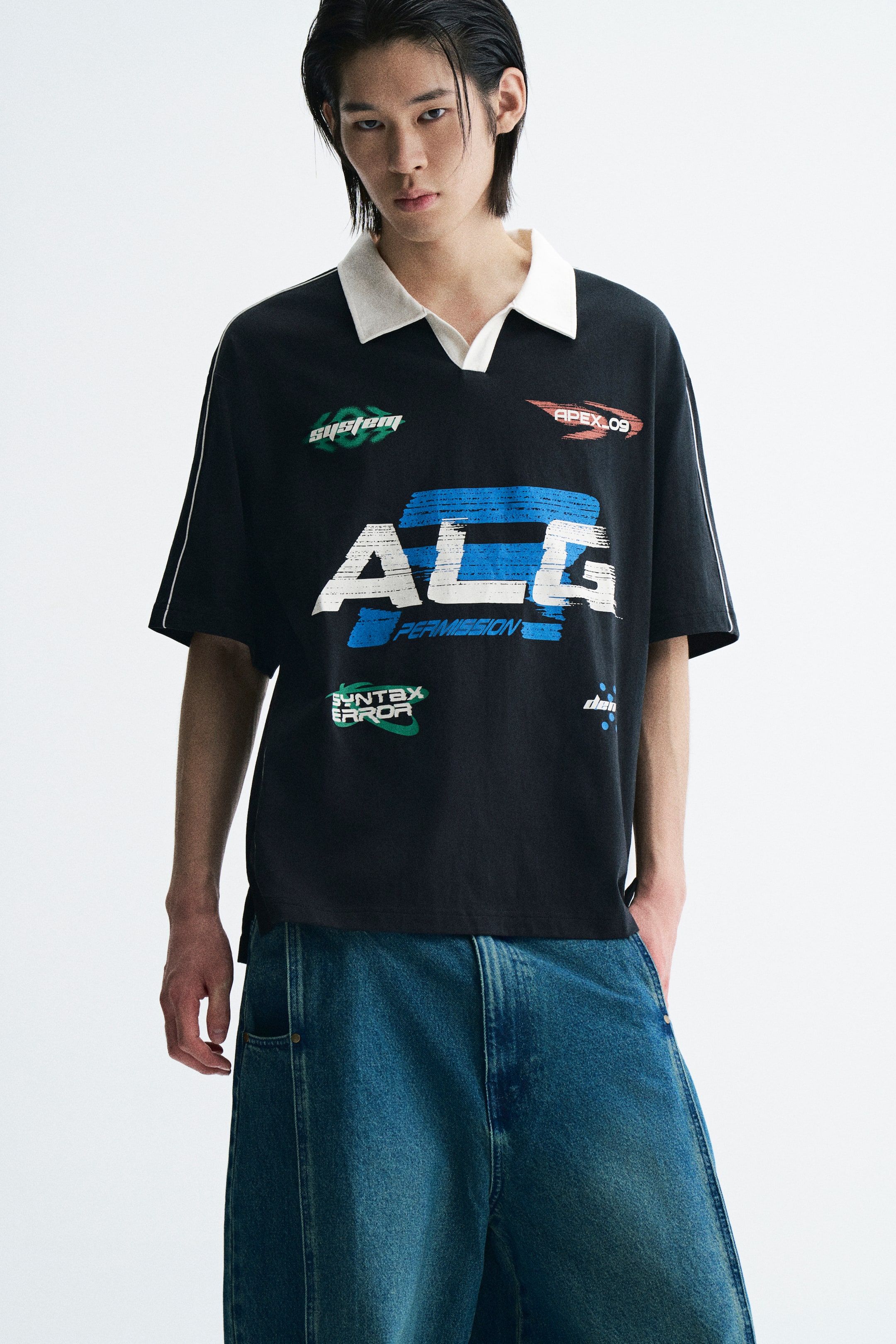 View larger image: The model is wearing a black, loose-fit short-sleeve polo shirt with a contrasting white open collar, dropped shoulders with white piping, and assorted graphic prints on the front. The shirt is styled with loose-fitting blue denim jeans.