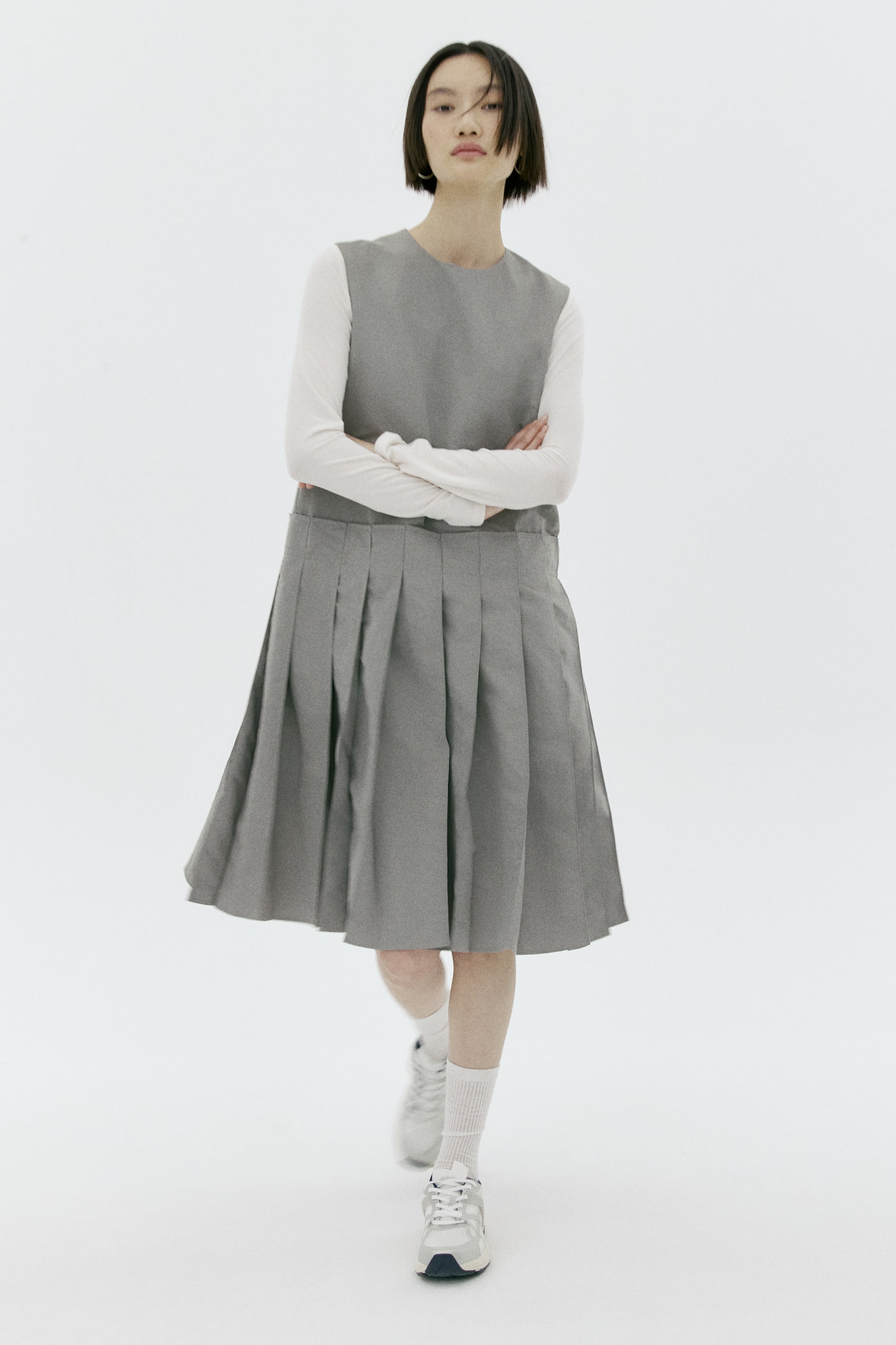 이미지 확대 보기: 회색 민소매 피나포어 원피스가 앞면에서 보이며, 라운드 네크라인과 선명한 나이프 플리츠(knife pleats) 처리된 스커트를 특징으로 합니다. 흰색 긴팔 톱 위에 레이어드되어 있습니다.