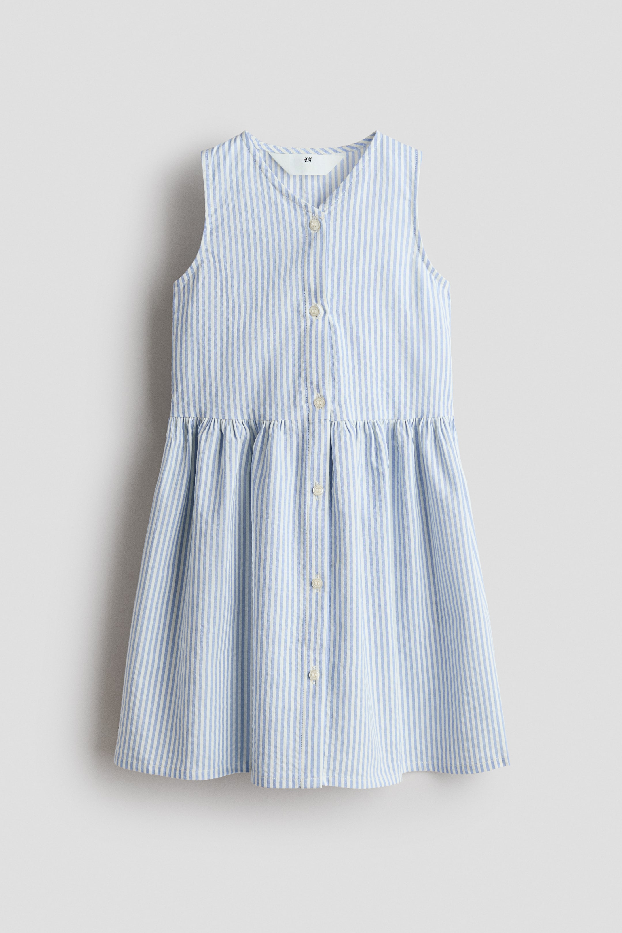 View larger image: The sleeveless dress showcases its front with a V-neck and buttons running down the front. Crafted from light blue and white striped seersucker fabric, it features a gathered seam at the waist, adding volume.