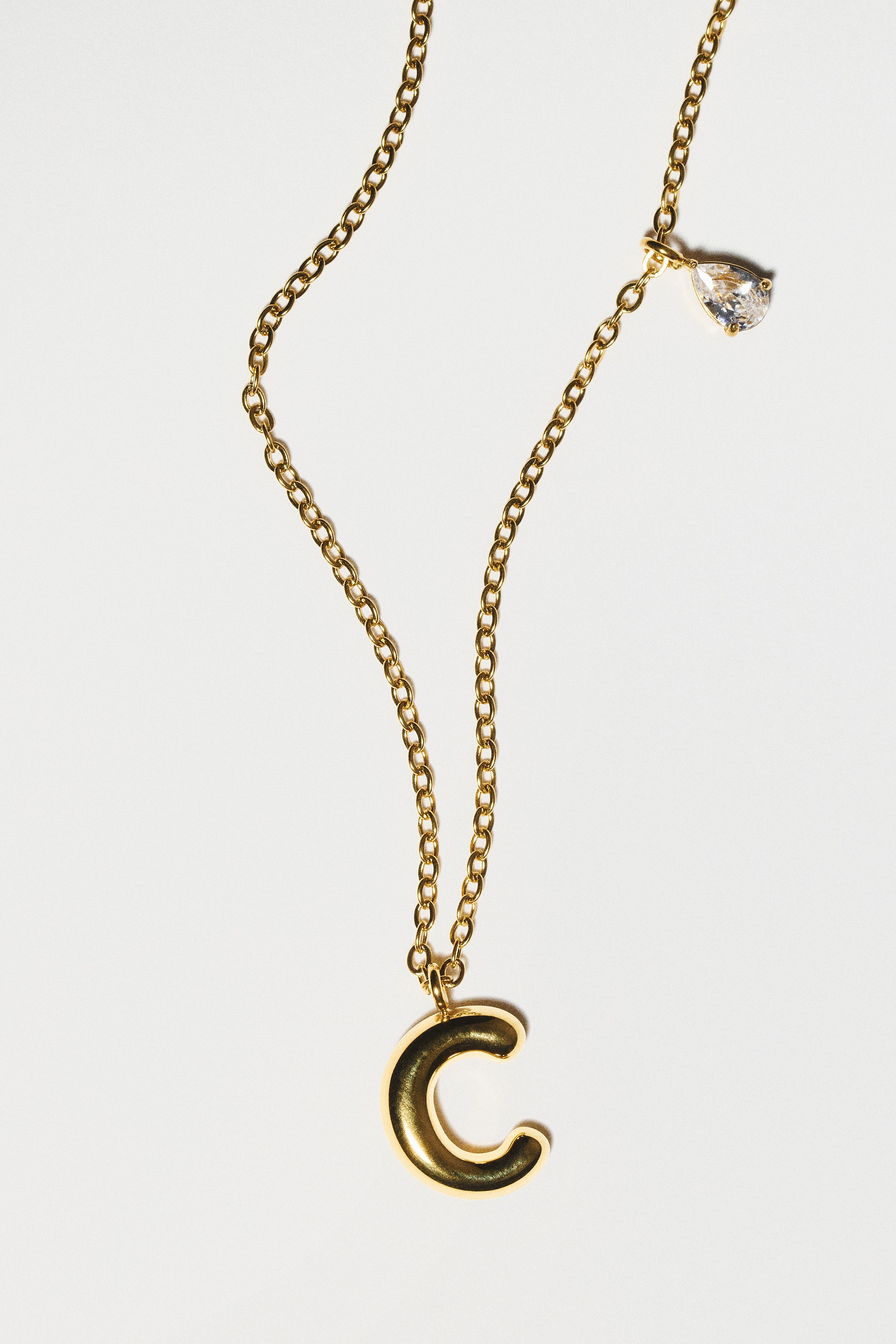 A gold-toned necklace features a fine link chain with a rounded, chunky initial 'C' pendant and a small, pear-cut clear crystal charm hanging at the centre, facing front.