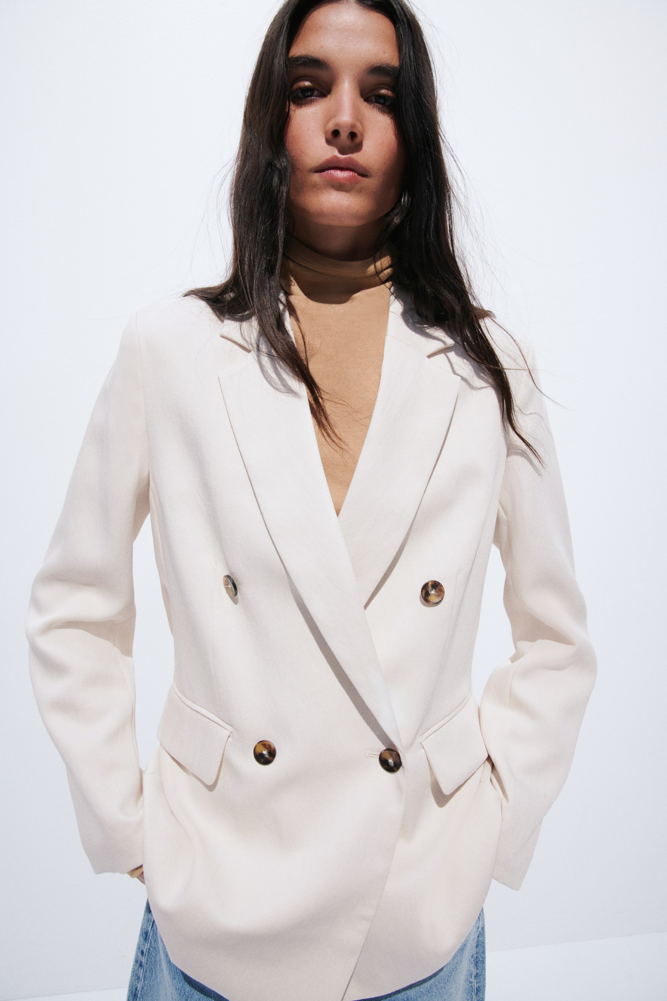 View larger image: A light cream double-breasted blazer featuring notch lapels, four visible tortoise-shell buttons, and two front flap pockets, worn open over a tan skivvy.