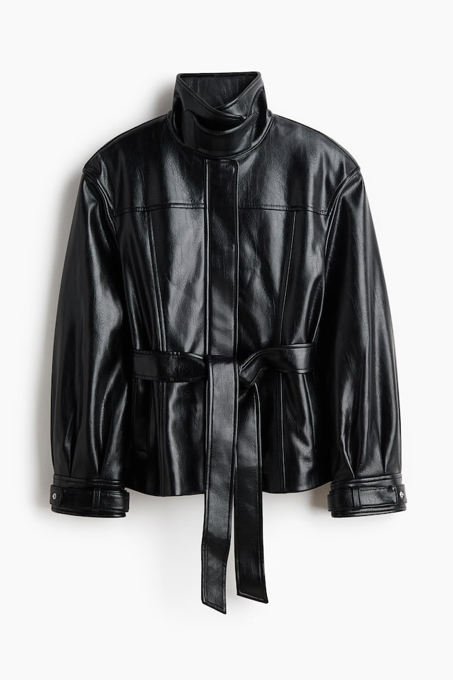 Une veste en similicuir noir est présentée de face, dotée d'un col montant, d'une patte de boutonnage cachée à l'avant et de coutures verticales définissant le torse. Une ceinture en tissu assorti serre la taille et se noue à l'avant, avec des manches longues se terminant par de larges poignets fixés par des boutons-pression argentés. - 1