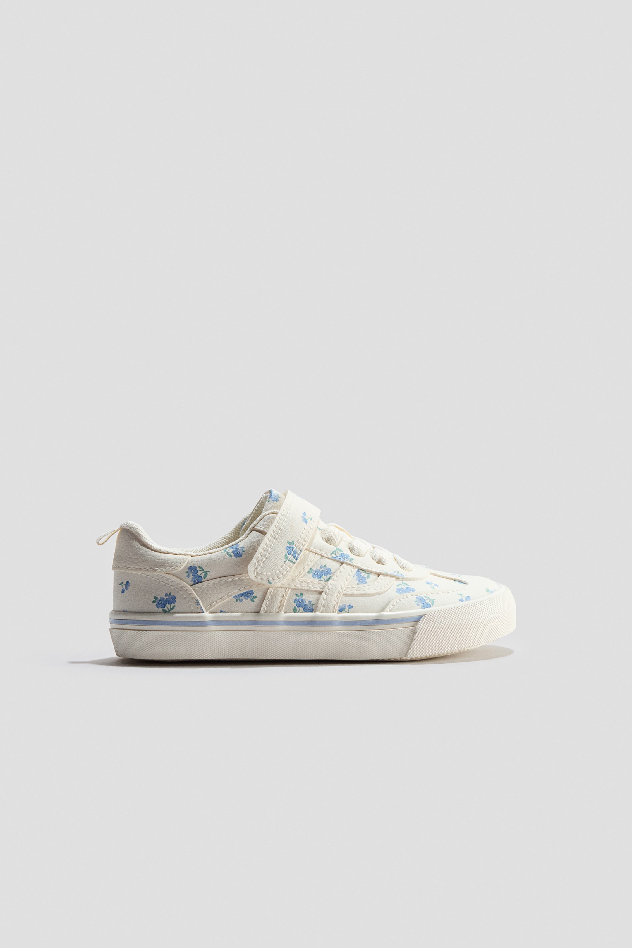View larger image: A cream canvas trainer is showcasing its side view, adorned with a blue floral pattern. It boasts a single Velcro strap fastening over the laces and a thick cream sole with a slim blue stripe.