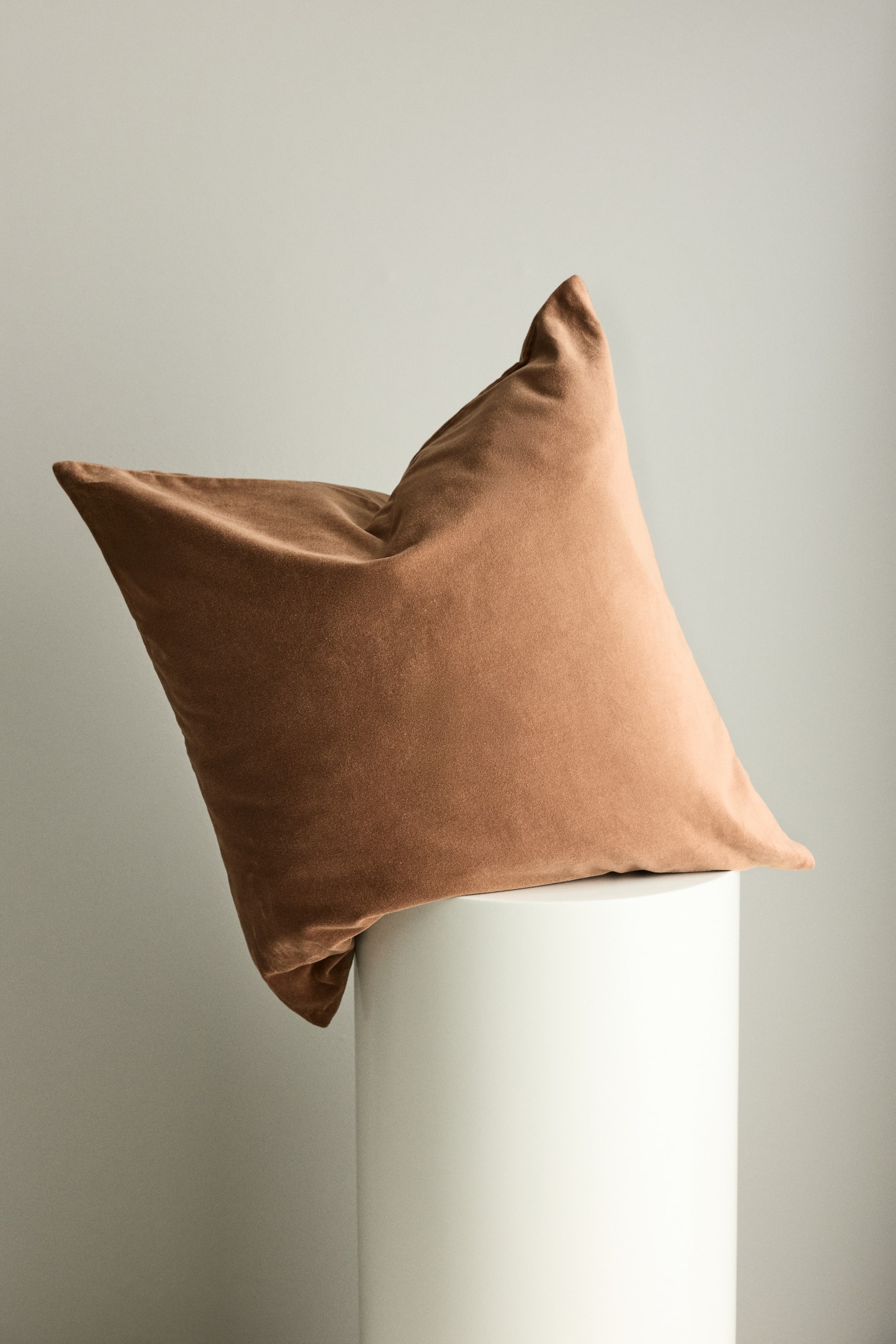 Visualizza immagine più grande: Un cuscino in velluto color cannella è appoggiato su un supporto cilindrico bianco, inclinato con l'angolo in alto a sinistra sollevato, mostrando la sua texture soffice e la superficie leggermente increspata.