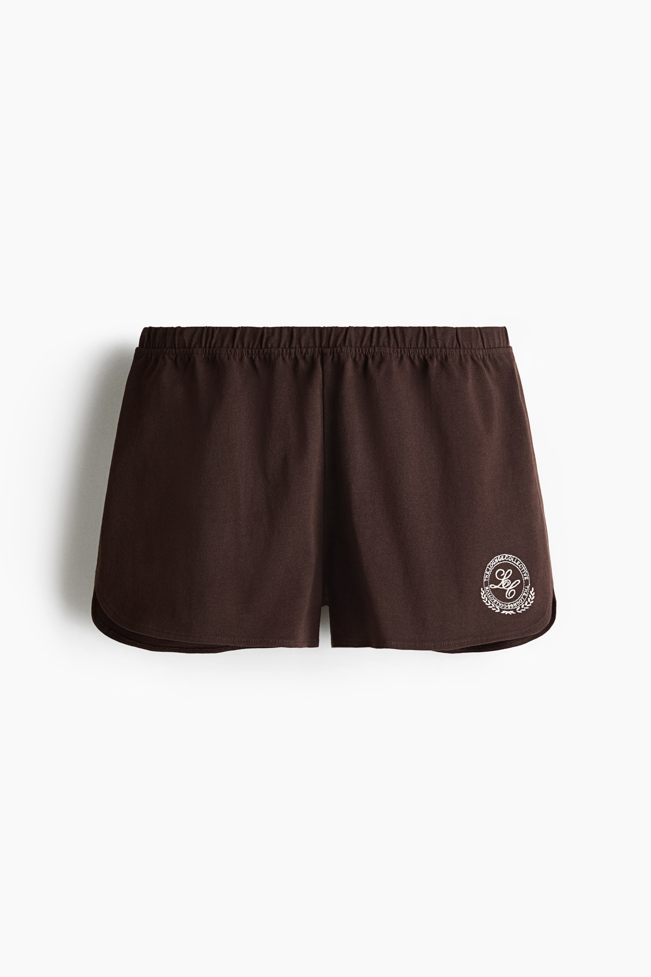 Grotere afbeelding bekijken: Deze donkerbruine katoenen jersey shorts worden van de voorkant getoond, met een bedekte elastische tailleband. Een cirkelvormig wit geborduurd motief met tekst en een krans is zichtbaar op de onderste rechterbeen.