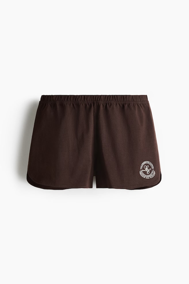 Estos pantalones cortos de jersey de algodón marrón oscuro se muestran desde el frente, con una cintura elástica cubierta. Un motivo circular bordado en blanco con texto y una corona es visible en la parte inferior de la pierna derecha. - 1