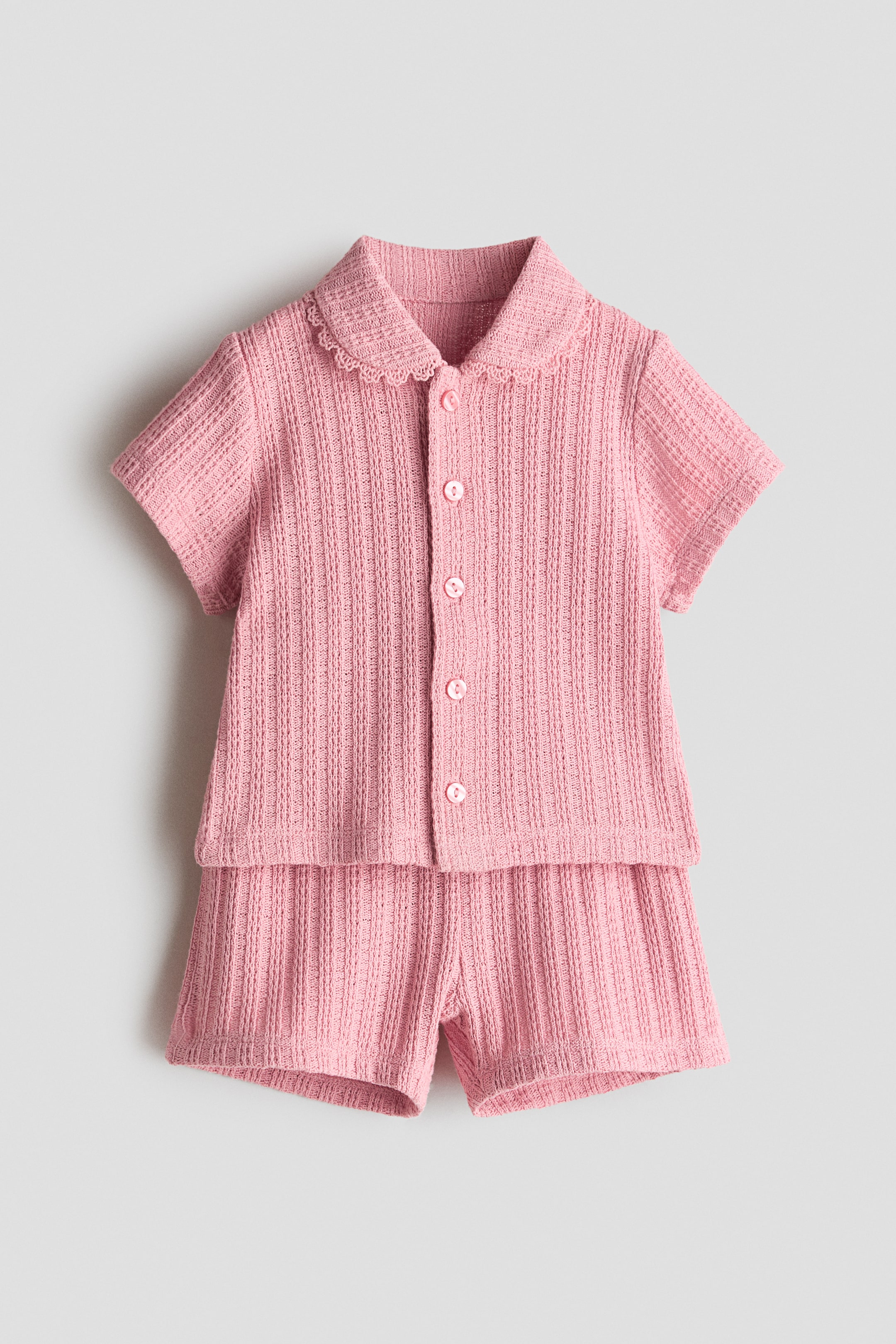 View larger image: A pink pointelle knit set is showcased at the forefront, comprising a short-sleeved shirt with a rounded, picot-trimmed collar and a front button placket, paired with matching shorts featuring an elasticated paper bag waist.