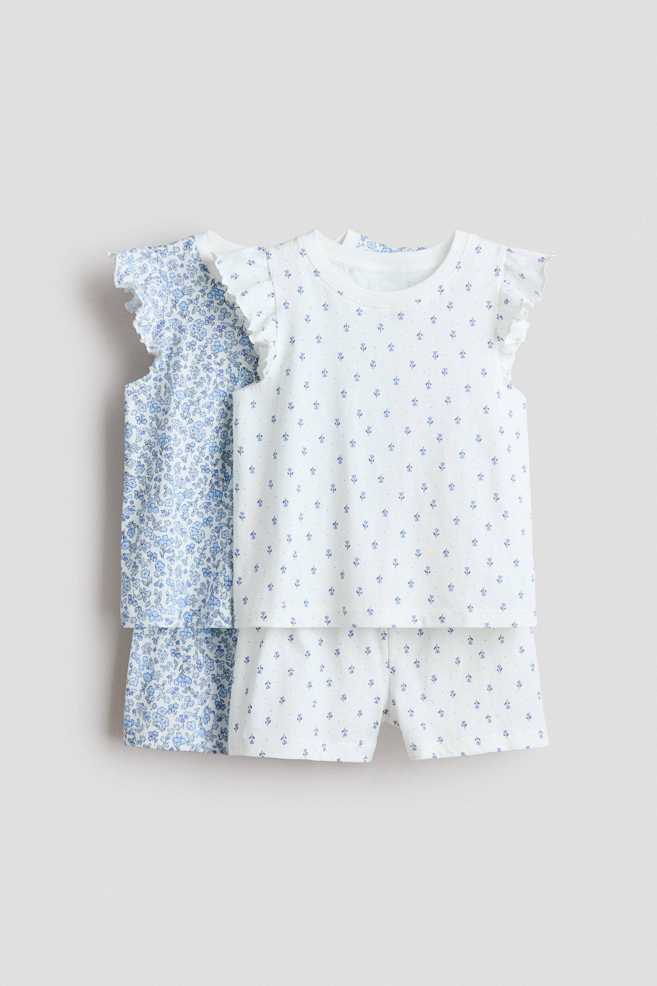 Grösseres Bild ansehen: Zwei Pyjama-Sets werden von vorne präsentiert, wobei das vordere Set ein weißes Oberteil und Shorts zeigt, beide mit einem Muster aus kleinen blauen Blumenmotiven und Punkten. Das weiße Oberteil ist mit einem runden, gerippten Ausschnitt und flatternden Ärmeln gestaltet, während ein ähnliches hellblaues Blumen-Set dahinter sichtbar ist.