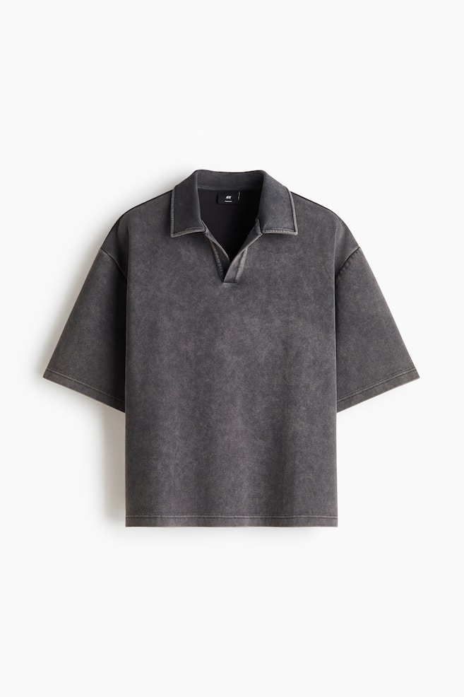 A short-sleeved polo shirt in a washed dark grey finish is facing front, showcasing its relaxed fit and dropped shoulders. It features a flat-lay collar with an open, buttonless V-neck placket and a straight hem. - 1