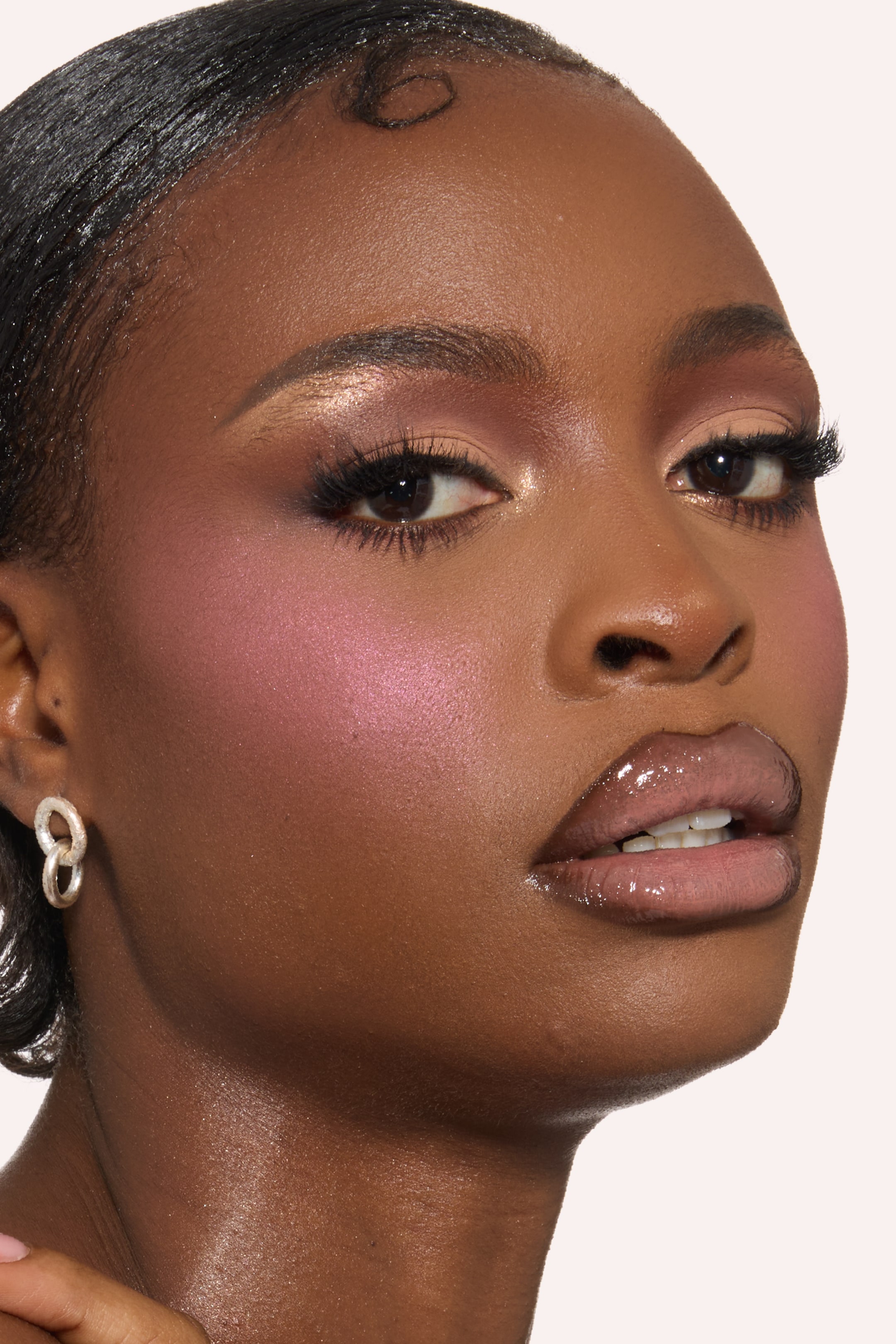 Visa större bild: Ett ansikte, lätt vinklat åt höger, visar upp en sminklook. Skimrande rosaskugga och långa mörka ögonfransar framhäver ögonen, medan ett lysande rosa rouge framhäver kindbenen. Glansig, nude läppstift definierar läpparna, och ett texturerat silvertonat dubbelhängsörhänge syns på vänster öra.