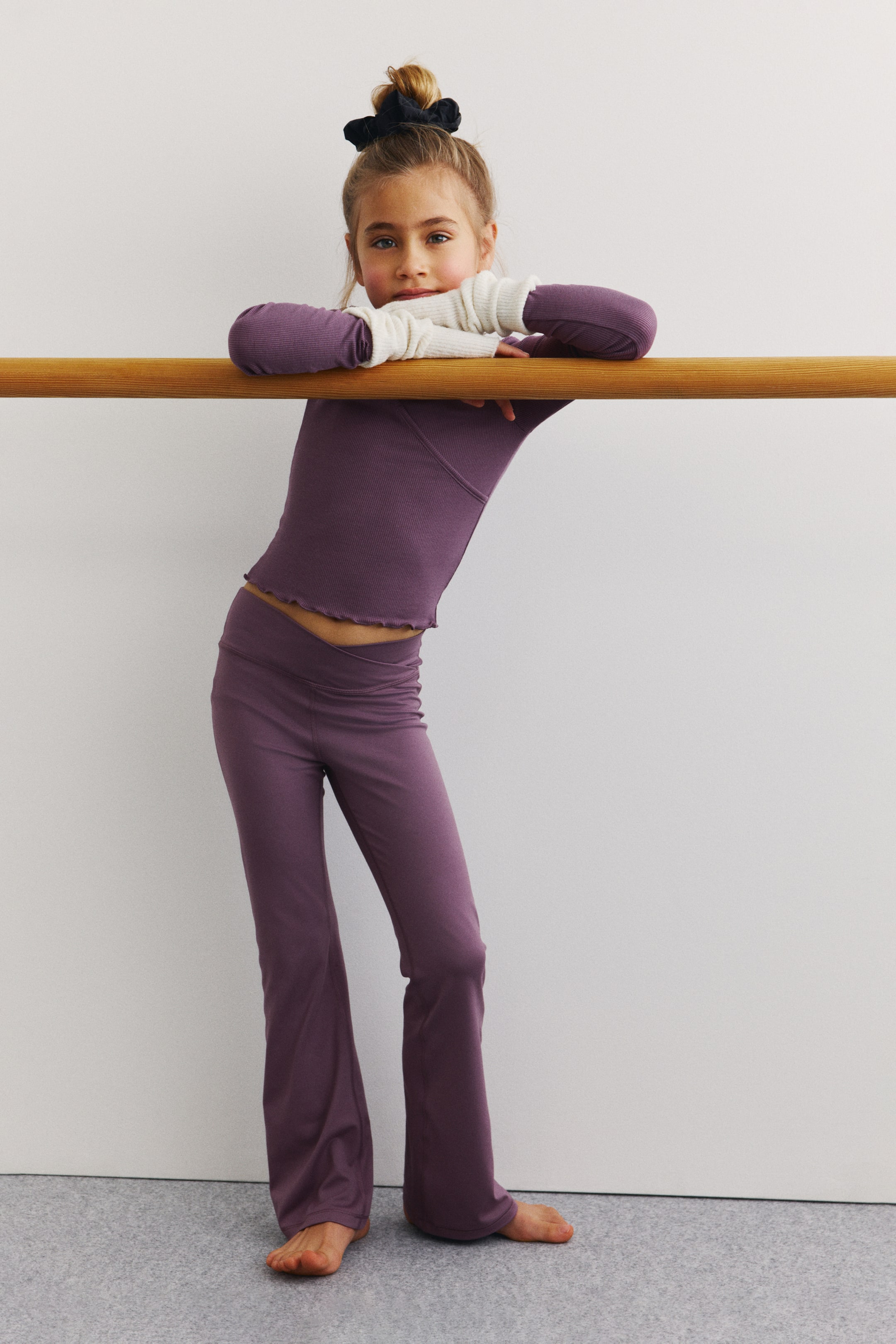 Ampliar la imagen: Un top morado de manga larga y punto acanalado, con dobladillo inferior tipo "lettuce-edge" y costuras en el pecho, se combina con pantalones acampanados morados a juego que presentan una pretina en forma de V. Manguitos blancos acanalados (arm warmers) se llevan en capas bajo las mangas del top, sobresaliendo más allá de los puños.