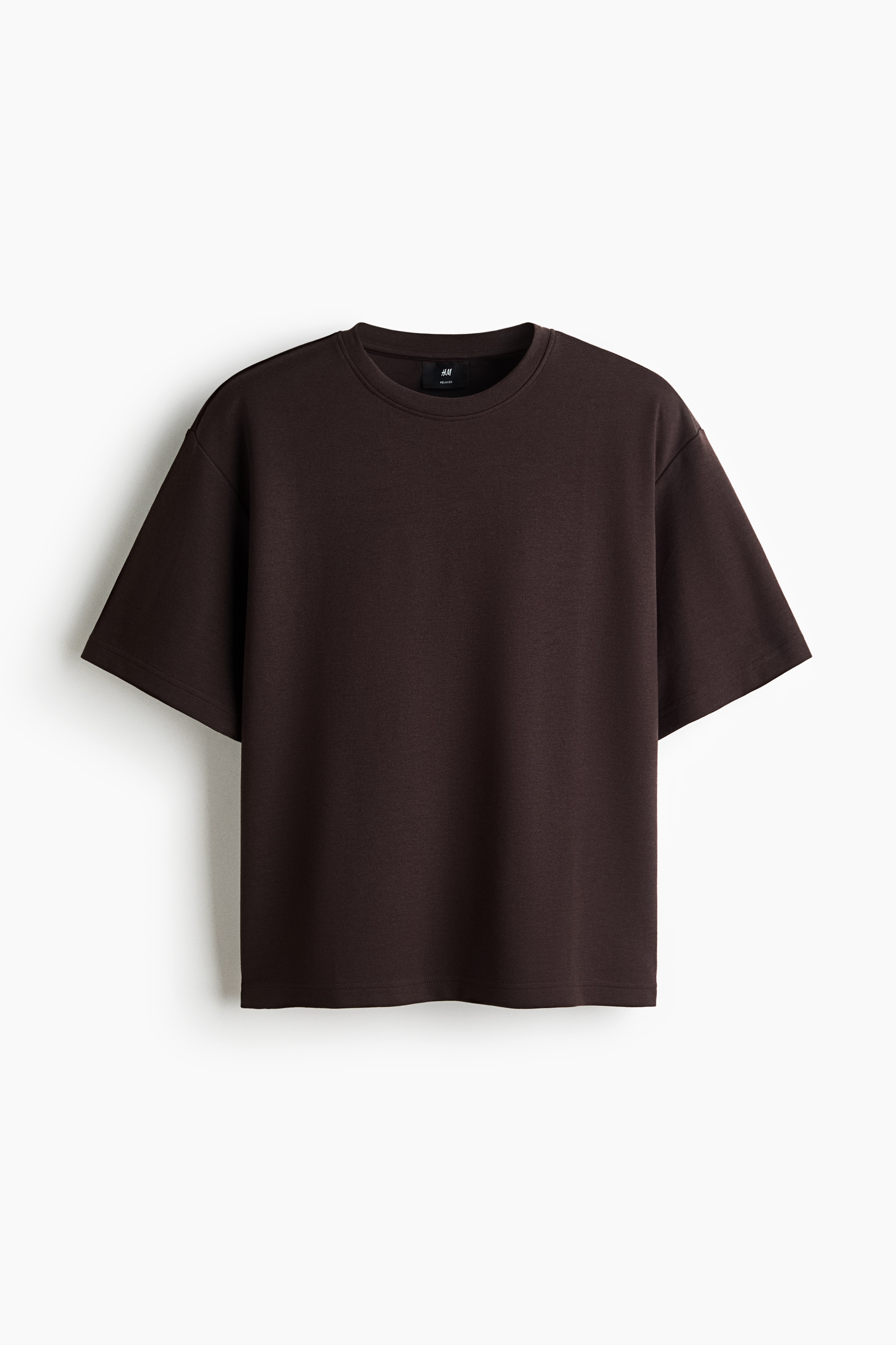 A dark brown heavyweight jersey T-shirt with a relaxed fit, round rib-trimmed neckline, and dropped shoulders is worn by the model. The model is standing with hands placed in the pockets of brown denim jeans.