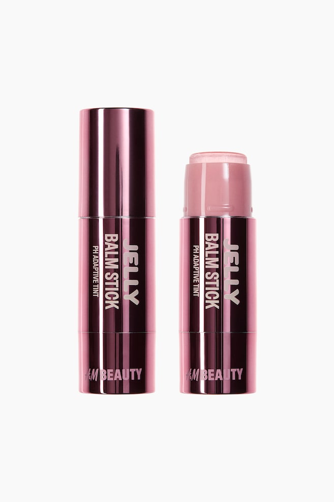 Two cylindrical balm sticks are standing upright, both featuring a metallic dark pink casing with white text on the FRONT. The stick on the left is closed, while the stick on the right is open, revealing a light pink, solid balm product extending from its top. - 1