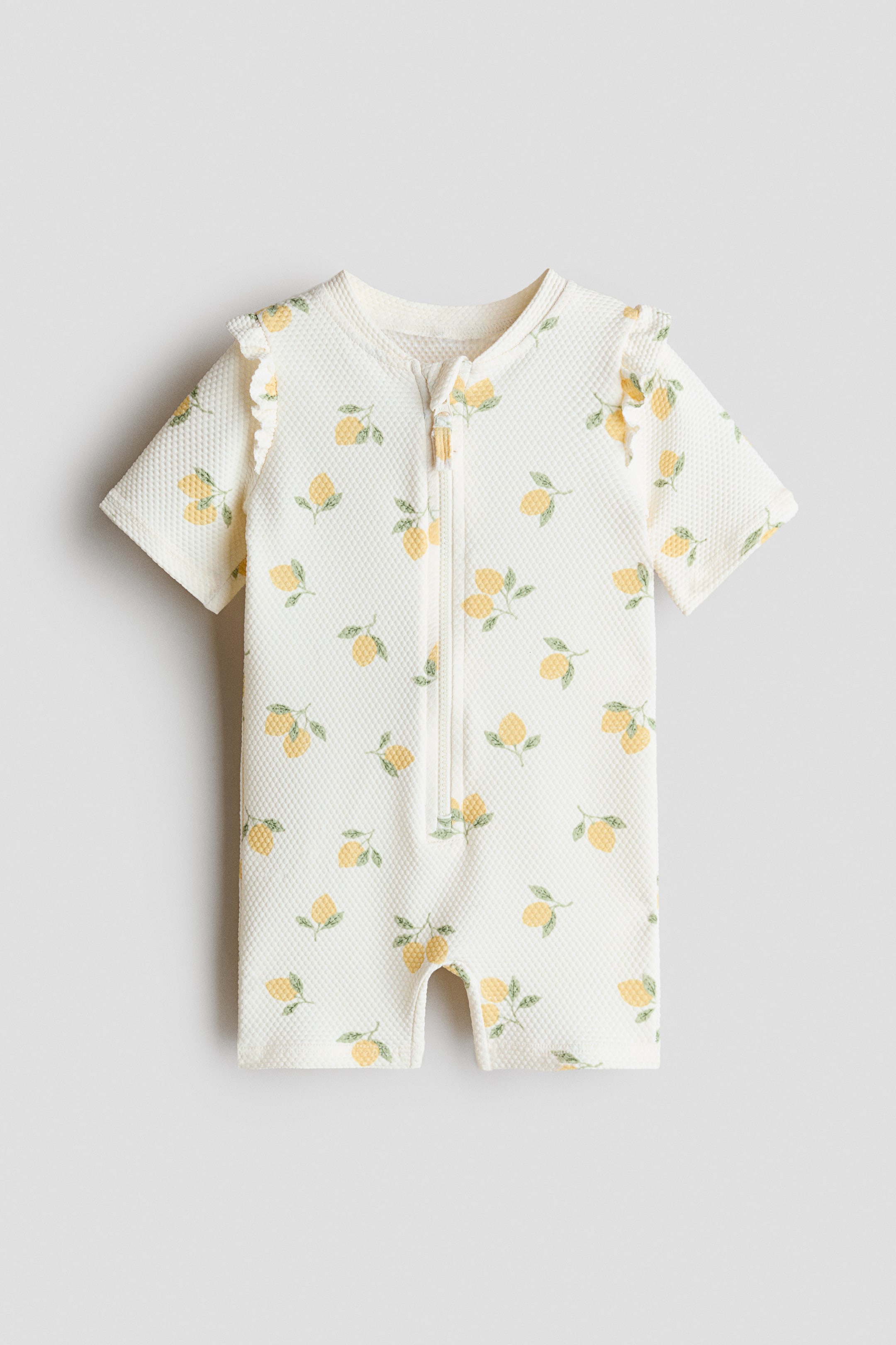 Zobacz większy obraz: Kostium kąpielowy w kolorze złamanej bieli z fakturą, skierowany przodem, ozdobiony powtarzającym się wzorem cytryn. Został zaprojektowany z zapięciem na zamek z przodu, krótkimi rękawami z falbanką na ramionach oraz krótkimi nogawkami.