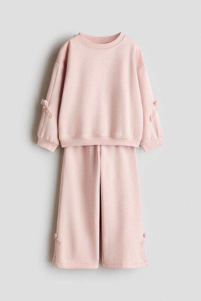 A light pink two-piece set is displayed front-facing, featuring a crew-neck sweatshirt and matching wide-leg pants. The sweatshirt shows ribbed trims and small decorative ribbon bows on the sleeves, while the pants feature matching ribbon details on the outer lower hems. - 1