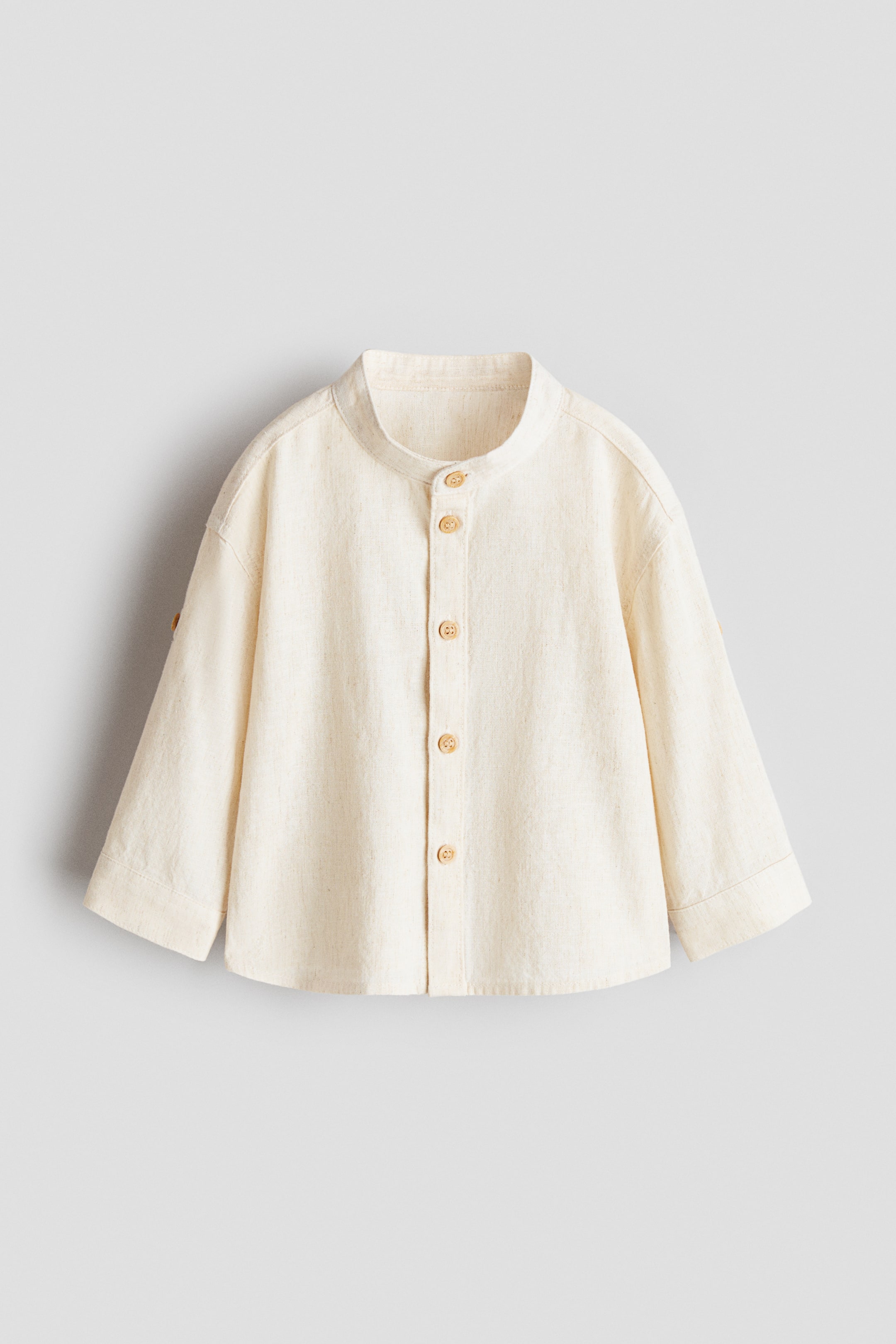 View larger image: Front view of an off-white shirt featuring a mandarin collar and a button placket with five light brown wooden buttons. The long sleeves are rolled up and secured with a buttoned tab.