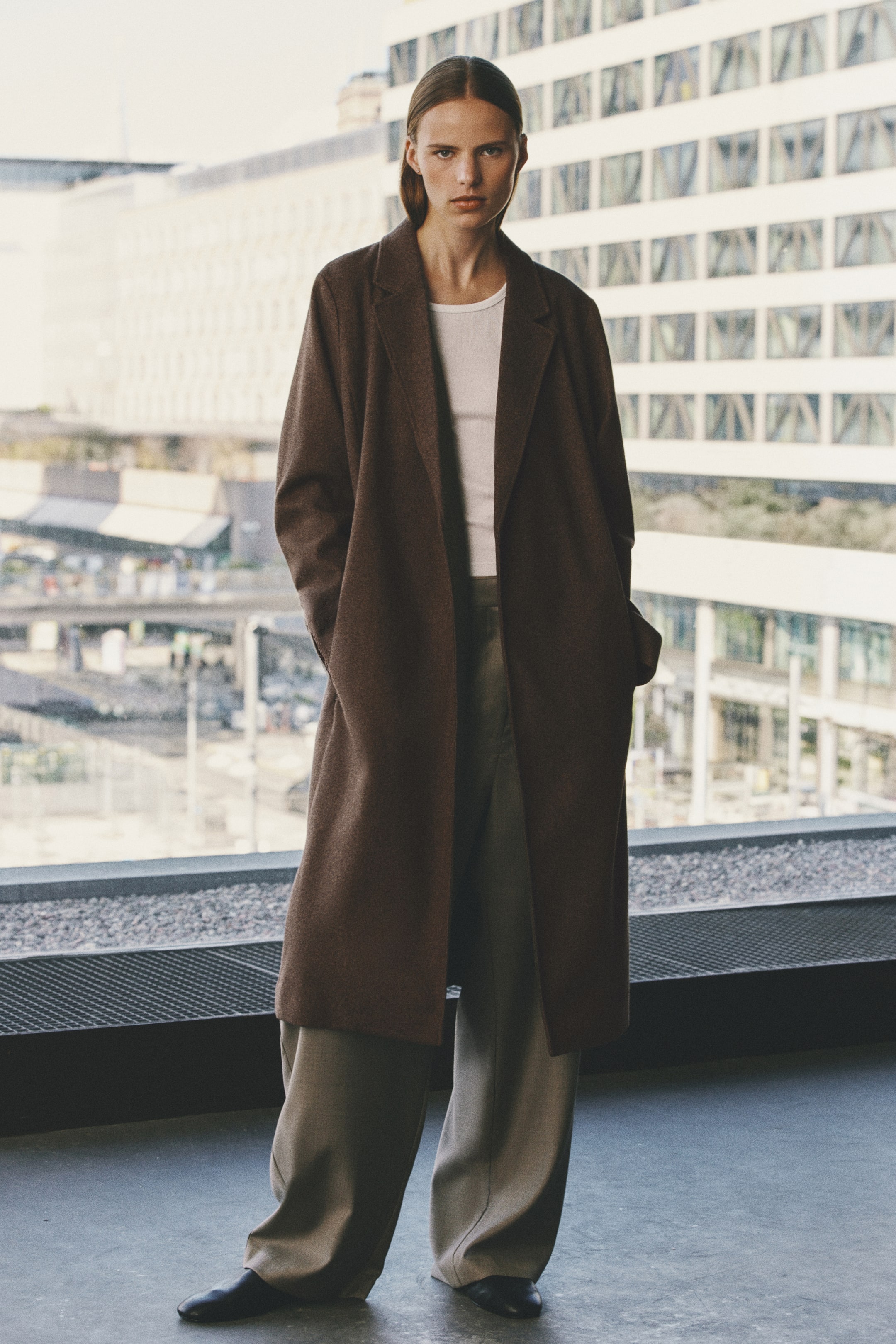 View larger image: The model is wearing a long brown coat in a brushed weave, worn open with both hands resting in the side pockets. The coat features prominent notch lapels and is styled over a white top and wide-leg trousers.