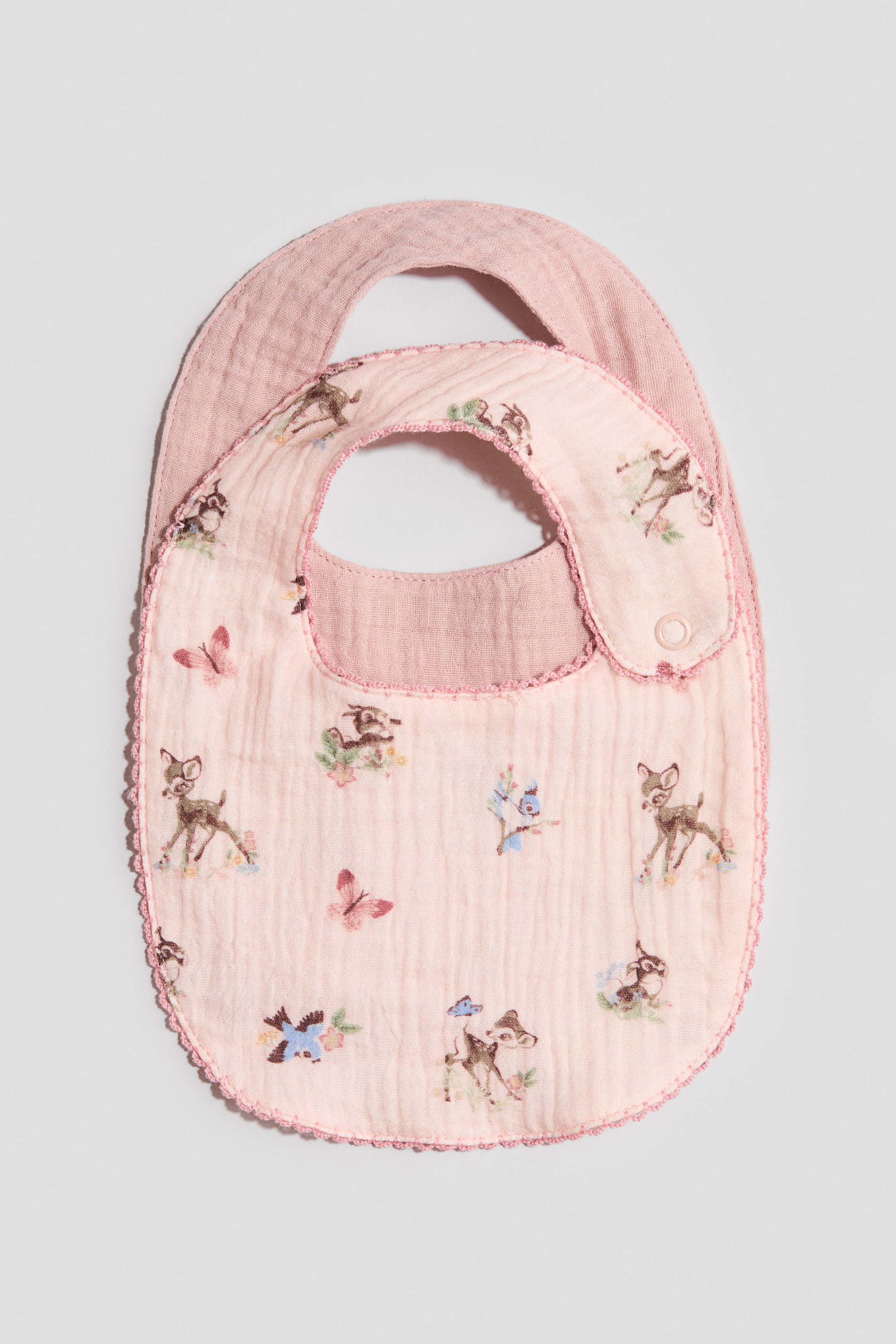 View larger image: Two baby bibs are presenting FRONT, layered with one slightly behind the other. The visible front bib is light pink and is featuring a repeating pattern of deer, butterflies, and flowers, with a decorative pink trim edging its perimeter and a single snap closure visible on the right shoulder strap; the bib layered behind it is a solid, slightly darker pink.