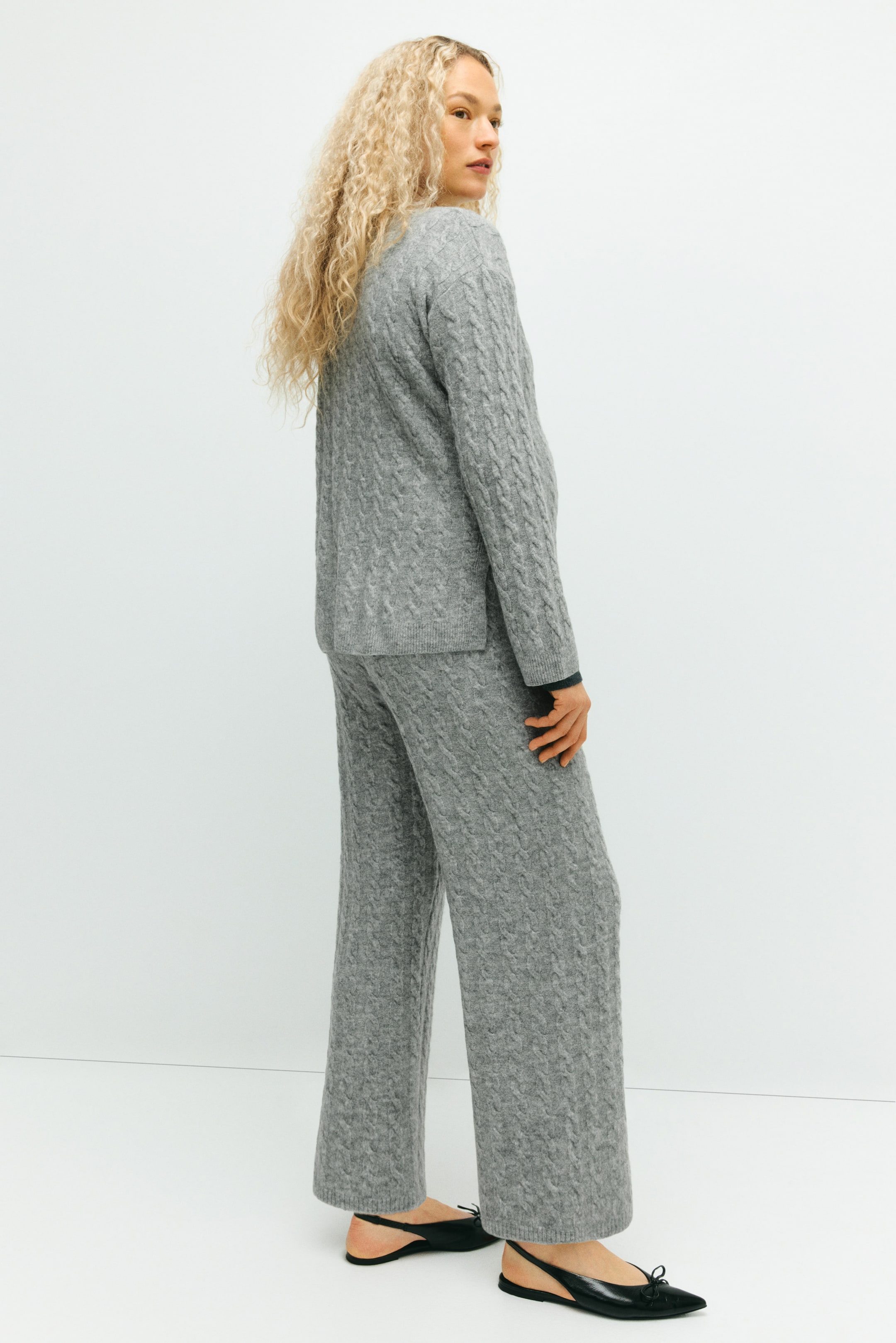 View larger image: A gray two-piece cable knit set is being presented from a 3/4 back angle, featuring a loose-fitting long-sleeved sweater and wide-leg pants. An intricate cable knit pattern is covering both pieces, with a dark long-sleeved garment layering beneath the sweater's cuffs.
