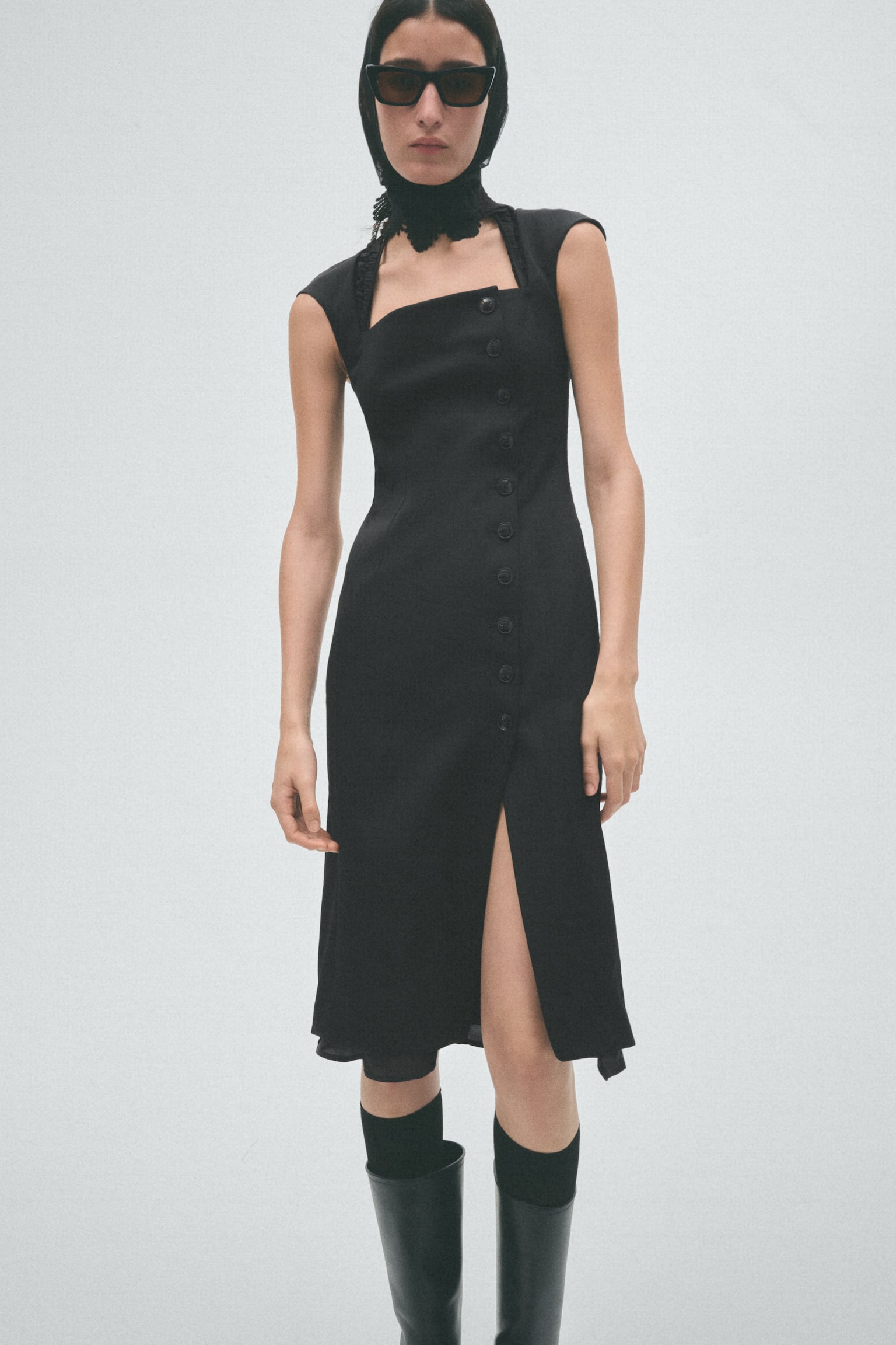 A black dress is hanging, facing forward, featuring a square bustline with structured cap sleeves and ruched detailing beneath the top edge, and a thin strap extending from the back neck. It is designed with an asymmetrical front placket, fastened by a row of black buttons, extending to a midi-length hem with a front slit. - 1