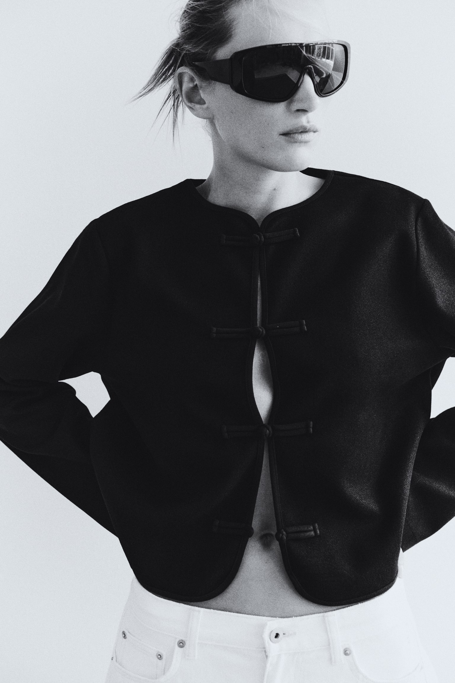 A black knit jacket with a round neckline, dropped shoulders, and front knot-button closures is being worn by the model. The jacket is styled partially open at the centre and paired with white trousers. - 1