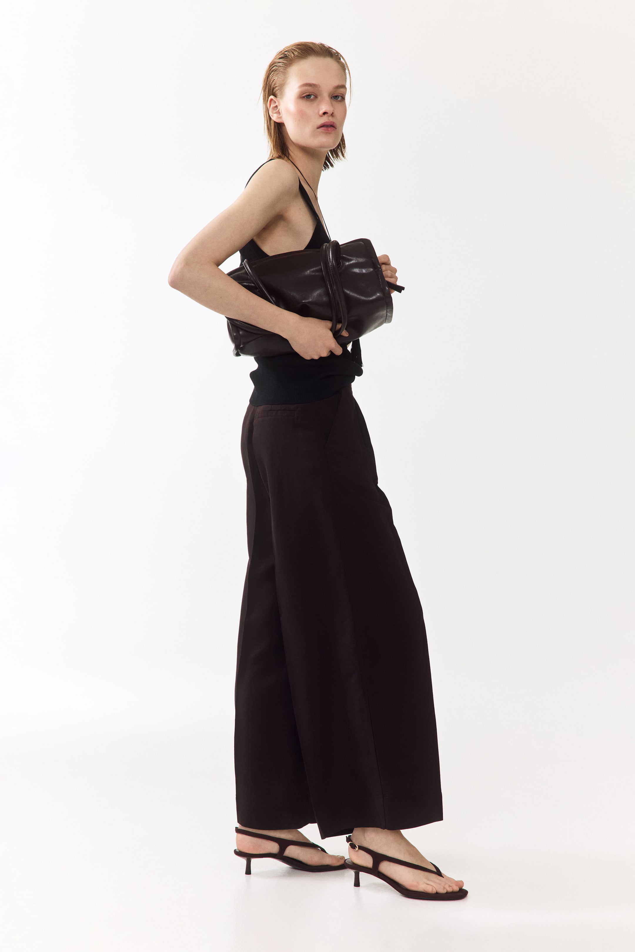 View larger image: A dark brown, soft leather handbag is being held by hands against the body from a side-front view, its elongated form featuring a gathered top closure and two soft top handles.