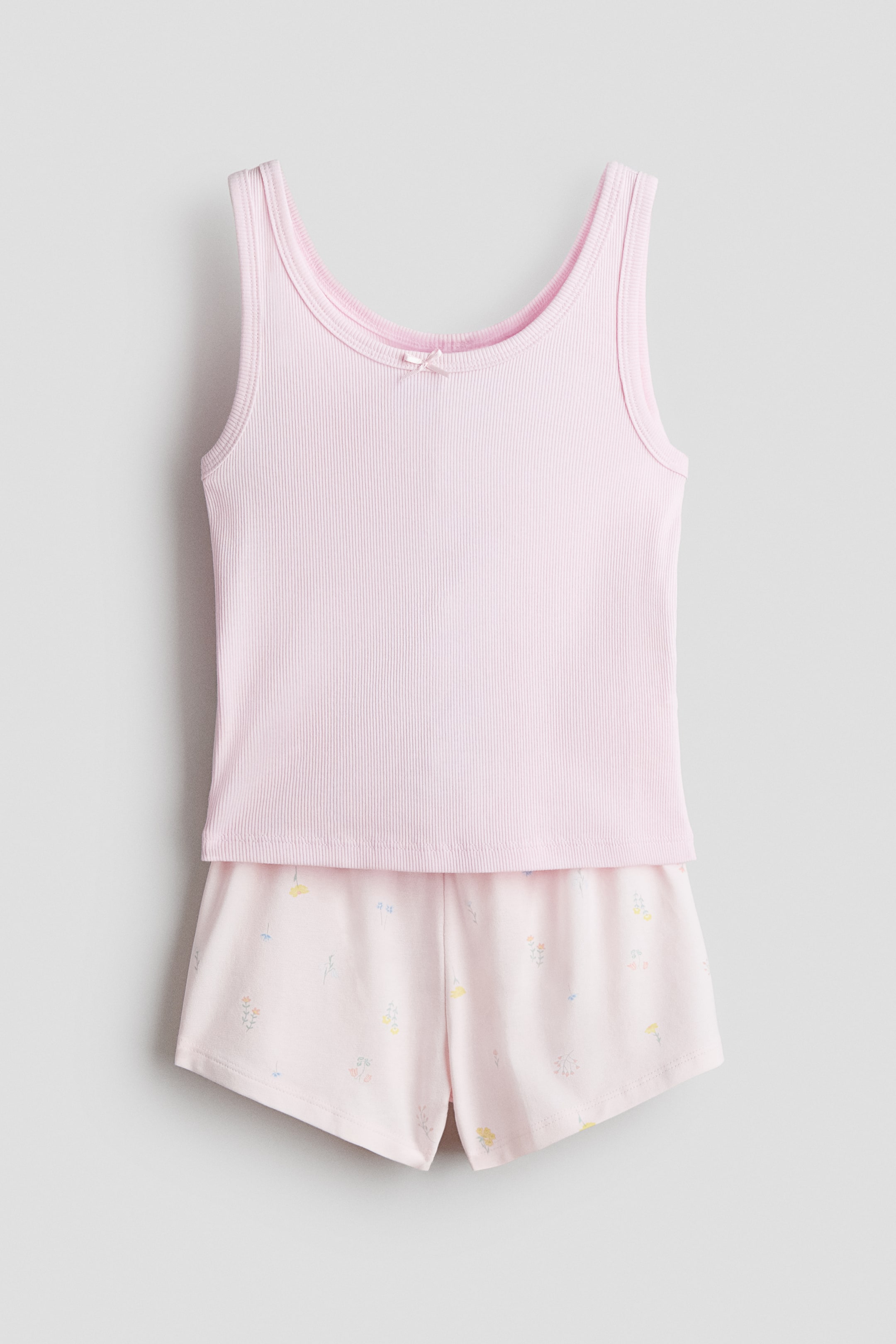 View larger image: A soft pink pajama set, comprising a ribbed tank top and shorts, is showcased front-facing. The tank top displays a scoop neckline adorned with a petite pink bow, while the shorts exhibit a graceful floral pattern with subtle botanical motifs in a variety of pastel hues.