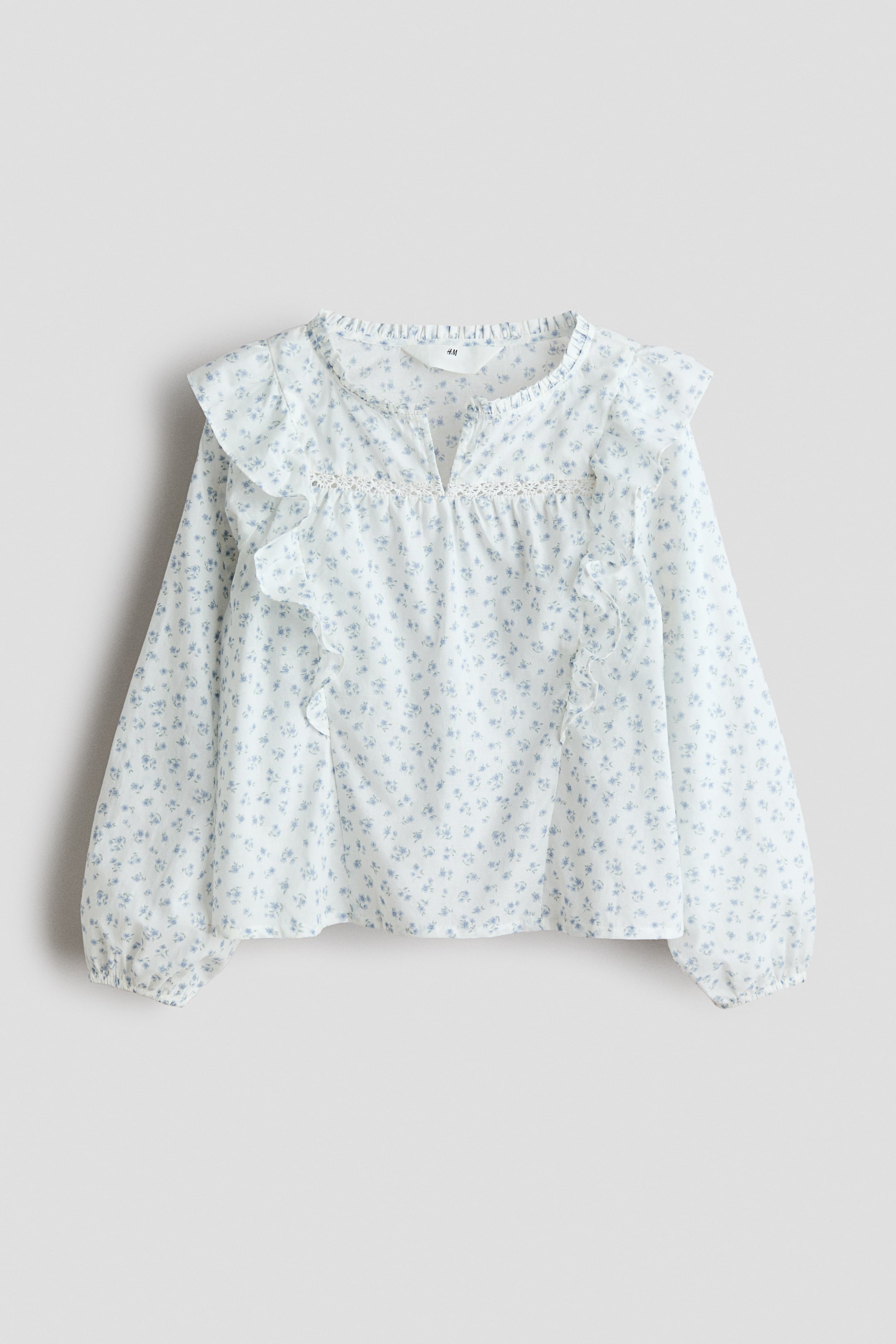 View larger image: A white blouse with a blue floral print is presenting front, featuring a gathered neckline with a V-split and white lace trim across the chest. Ruffles are extending from the neckline over the shoulders and down the front, and the long sleeves are gathering at the cuffs.