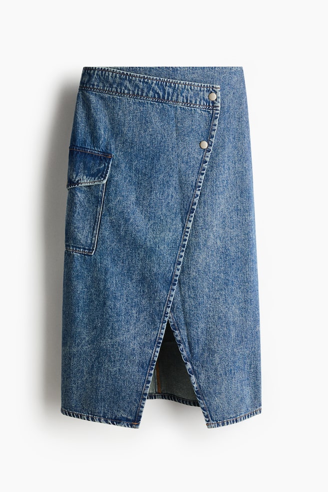 A blue denim wrap skirt is facing front, featuring an asymmetrical front panel that overlaps and secures with two silver-tone buttons. The mid-wash denim skirt is displaying a side utility flap pocket and a front slit created by the wrap design. - 1
