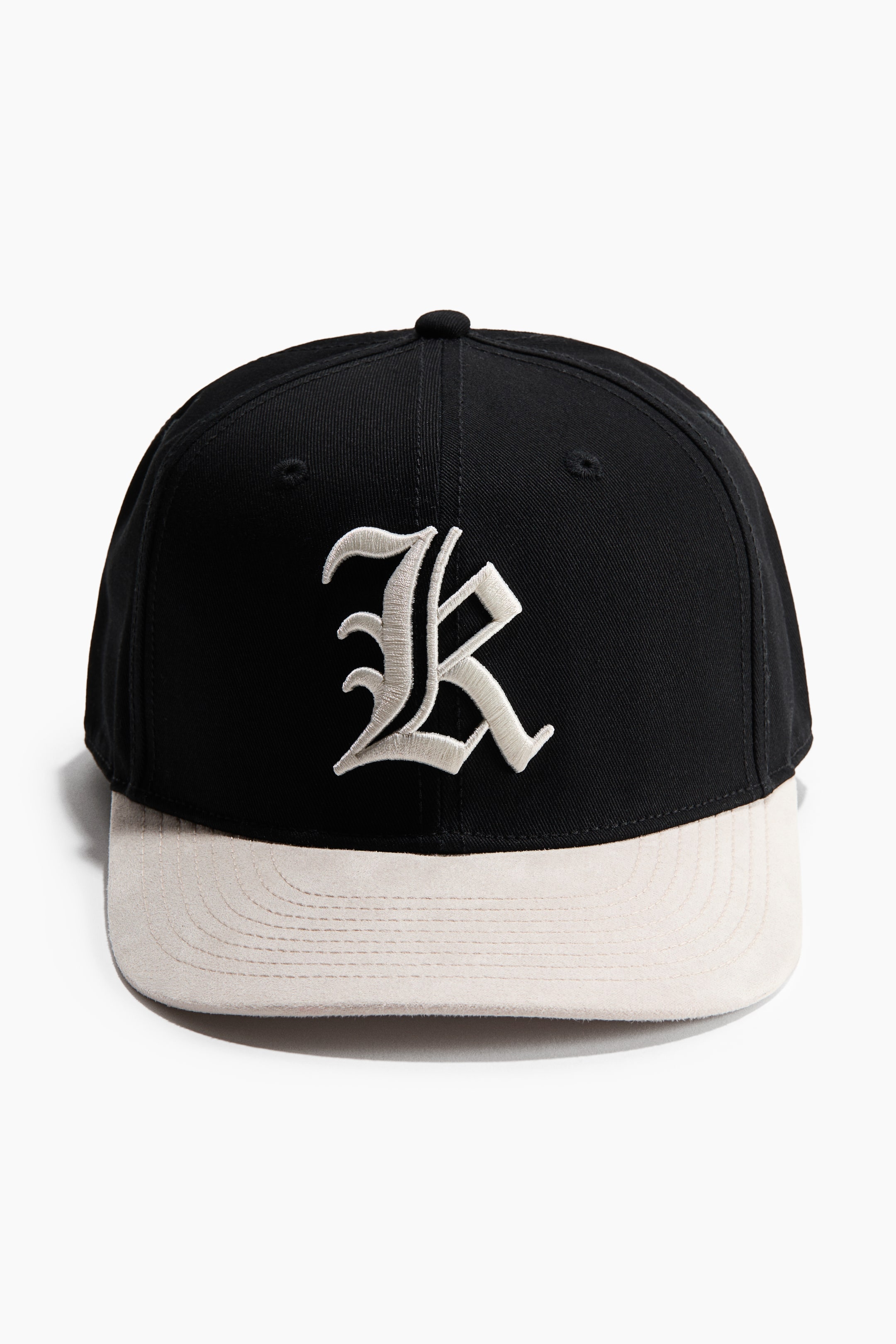 View larger image: The cap is oriented forward, showcasing a black twill crown paired with a light beige flat brim. Prominently featured on the front is a large, stylized letter R in light beige embroidery, with embroidered eyelets visible on the crown.