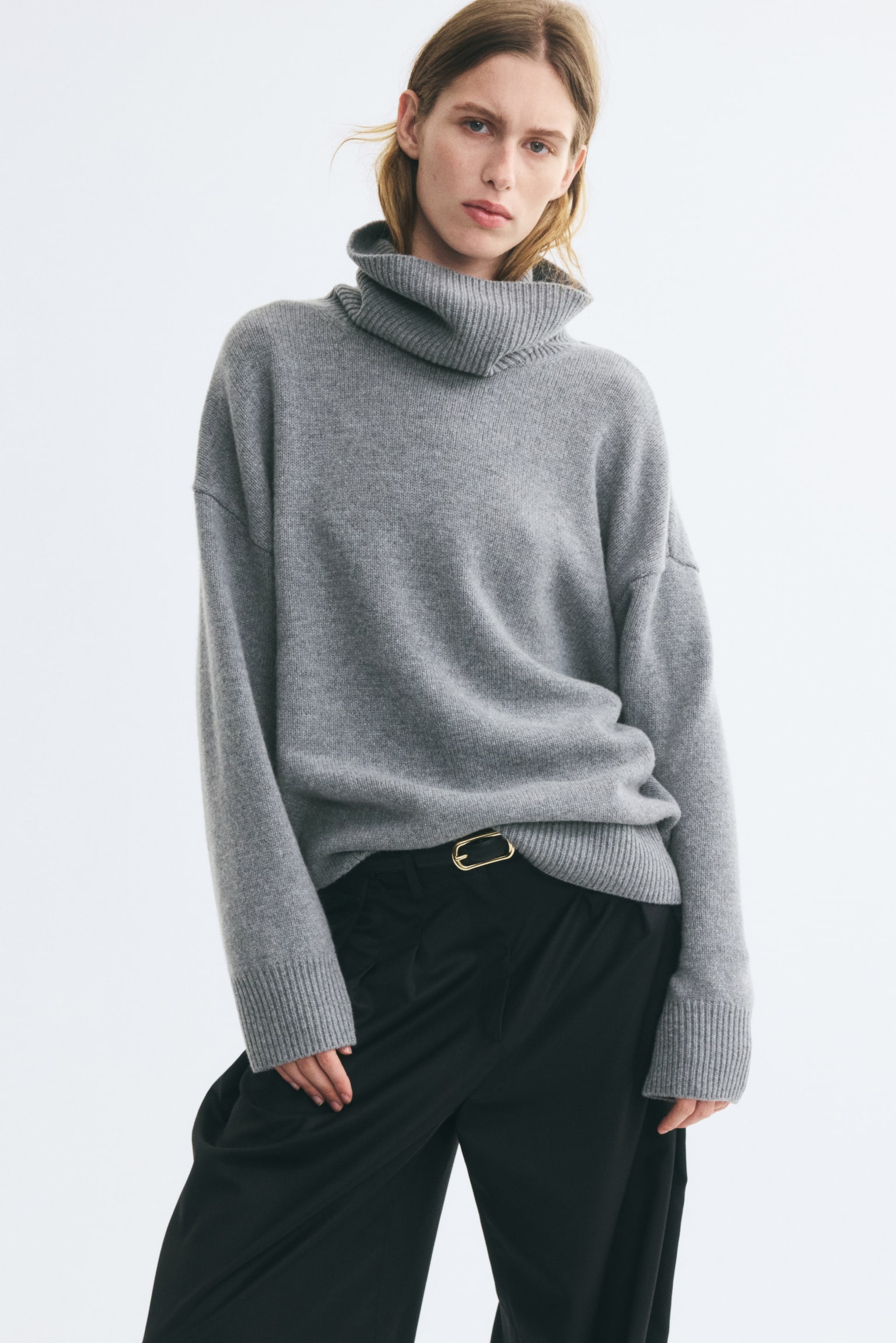 An oversized grey knit sweater featuring a ribbed turtleneck, dropped shoulders, and wide ribbed cuffs is worn by the model. The hem of the sweater is tucked into black trousers styled with a gold-buckled belt. - 1