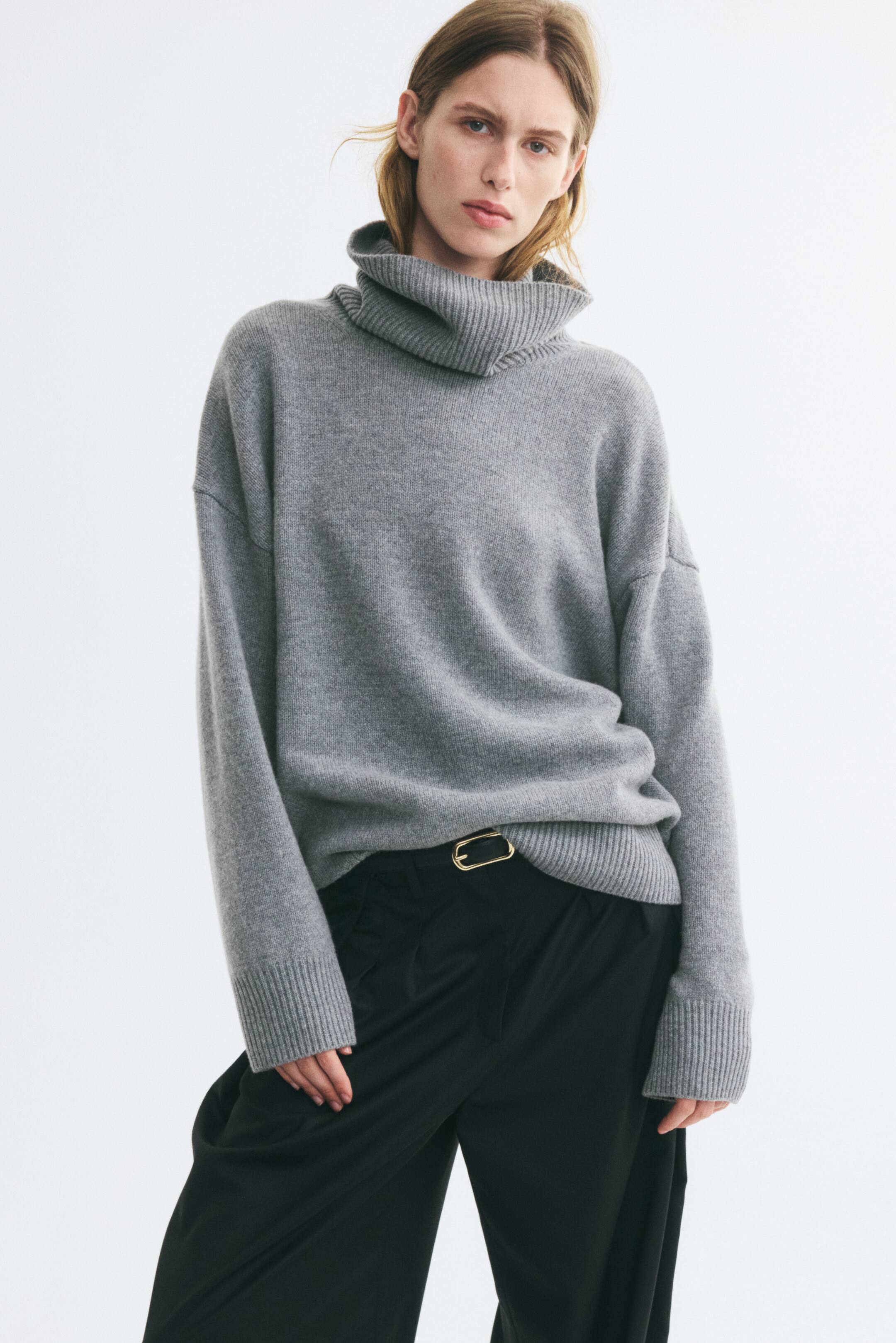 Προβολή μεγαλύτερης εικόνας: Ένα oversized γκρι πλεκτό πουλόβερ με ριμπ ζιβάγκο, χαμηλωμένους ώμους και φαρδιές ριμπ μανσέτες φοριέται από το μοντέλο. Το τελείωμα του πουλόβερ είναι χωμένο μέσα σε μαύρο παντελόνι, σε συνδυασμό με ζώνη με χρυσή αγκράφα.