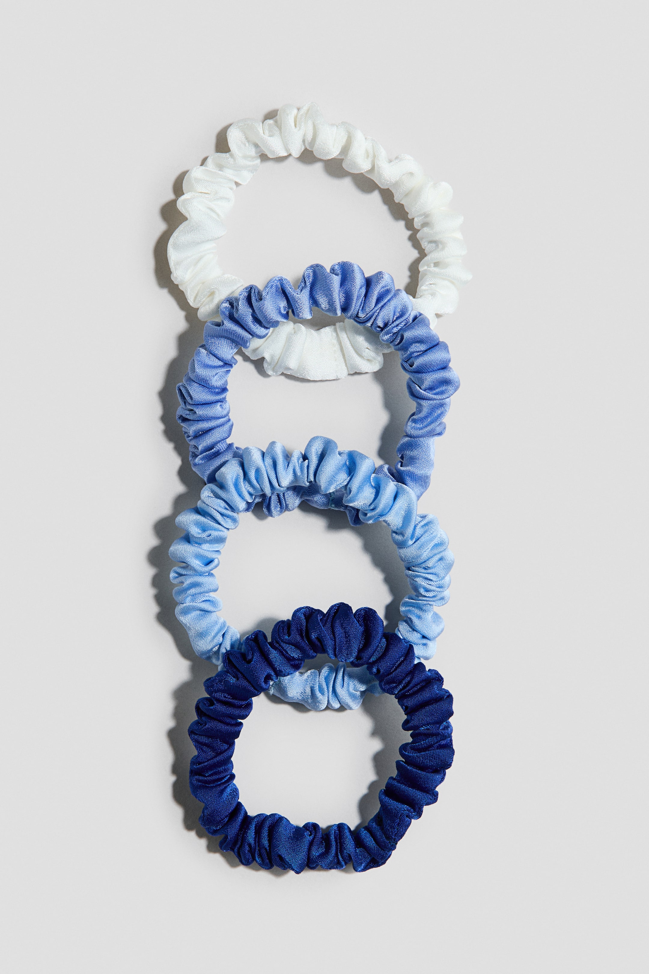 Se større billede: Et sæt med fem scrunchies er arrangeret lodret og viser en farvegradient fra hvid til dyb marineblå. Hver scrunchie har en samlet, rynket tekstur og en diskret glans.