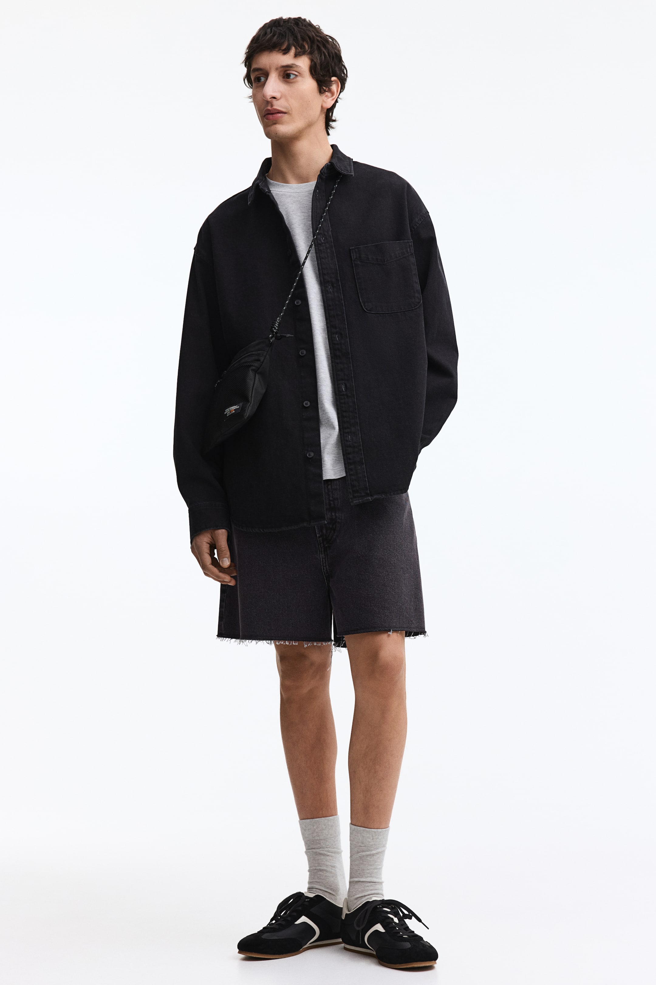 View larger image: A black denim overshirt is displayed from a front-facing angle, worn open over a light gray t-shirt. It features a spread collar, a button-up front closure, a single chest patch pocket, and a frayed bottom hem.